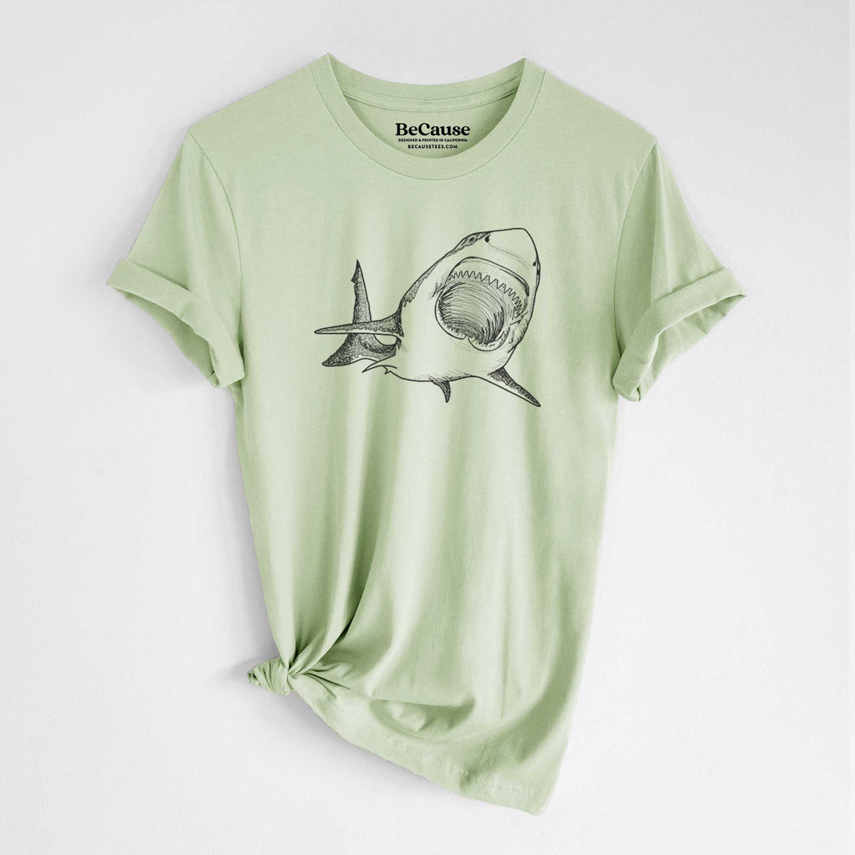 Spring green lightweight cotton t-shirt laid flat with the sleeves rolled, printed with a black ink illustration of a great white shark swimming toward the viewer with its mouth open.