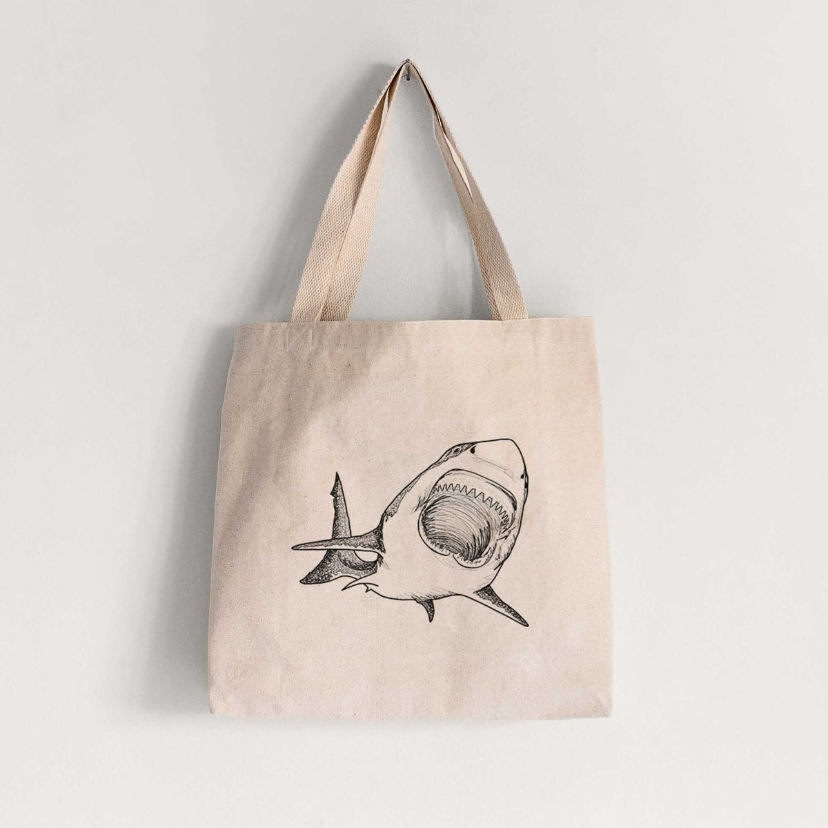 Natural canvas tote bag printed with a black ink illustration of a great white shark swimming toward the viewer with its mouth open.