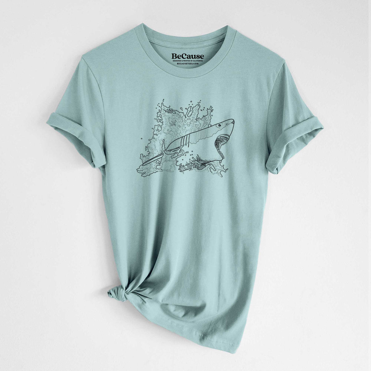 Dusty blue lightweight cotton t-shirt laid flat with the sleeves rolled, printed with a black ink illustration of a great white shark breaking through a splash of water.