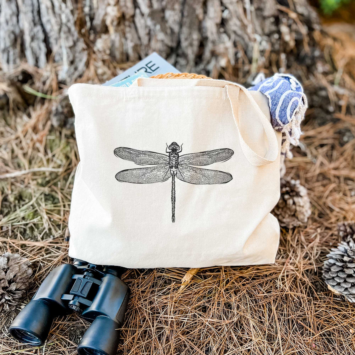 Canvas tote bag printed with a detailed black ink illustration of a green darner dragonfly with its wings spread, seen from above, resting on dry grass beside a pair of binoculars.
