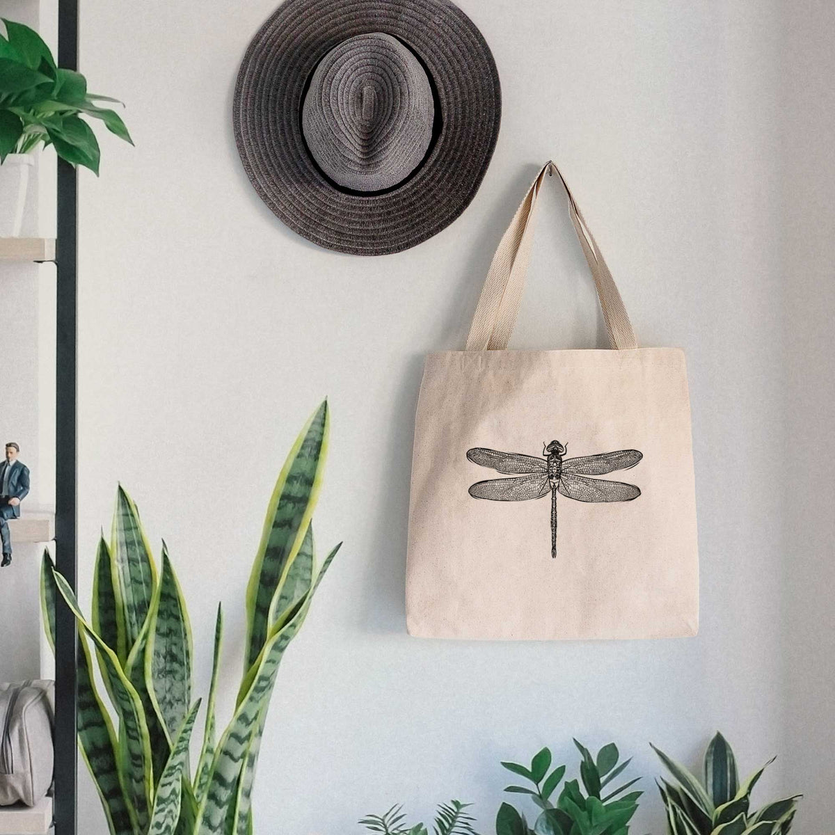 Canvas tote bag printed with a detailed black ink illustration of a green darner dragonfly with its wings spread, seen from above, hanging on a wall hook below a hat, next to potted plants.