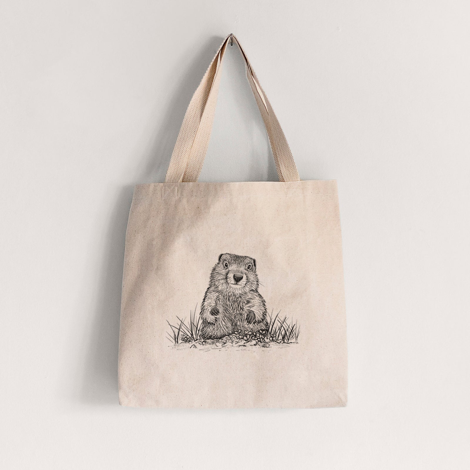 Natural canvas tote bag printed with a black ink illustration of a groundhog peeking out of its burrow among grass.