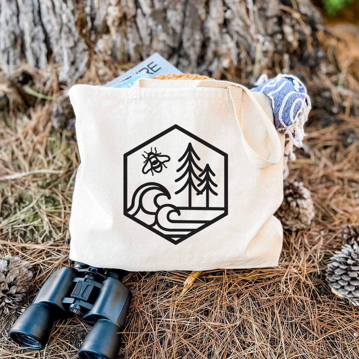 Canvas tote bag printed with a black line art hexagon holding a bee, ocean waves and pine trees, resting on dry grass beside a pair of binoculars.