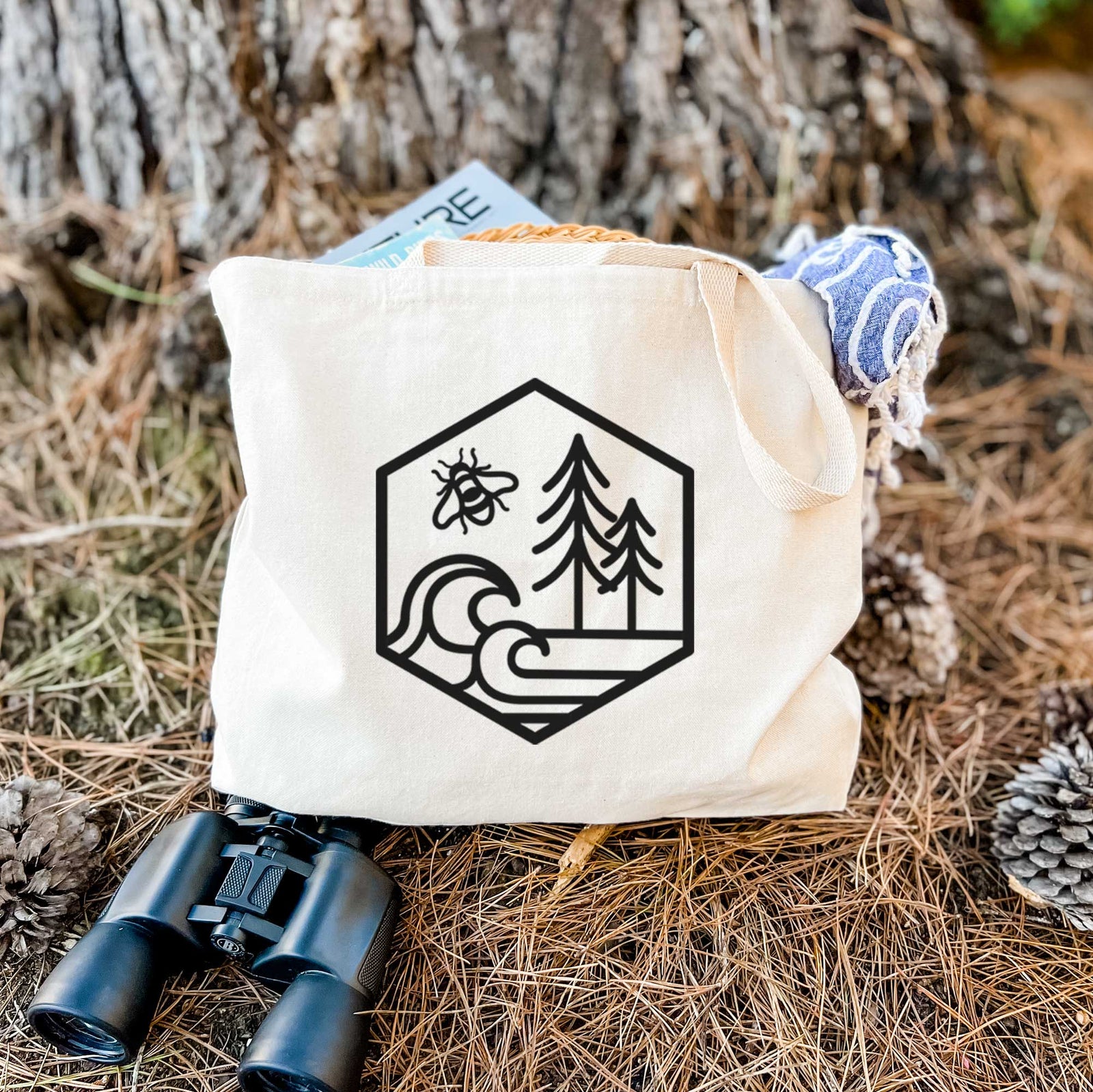 Natural canvas tote bag printed with a black line art hexagon holding a bee, ocean waves and pine trees.