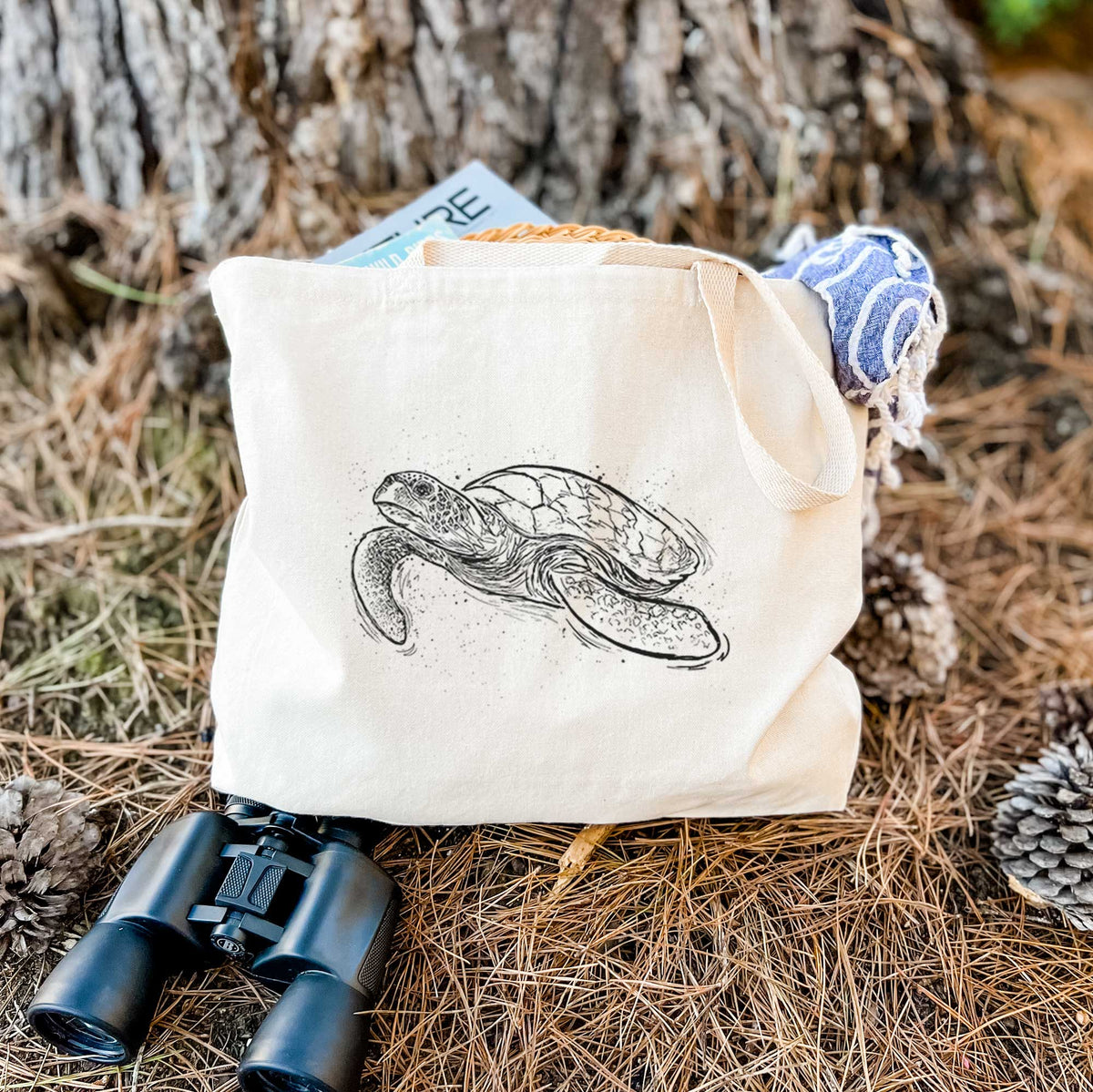 Canvas tote bag printed with a black ink illustration of a hawksbill sea turtle swimming, with a patterned shell, resting on dry grass beside a pair of binoculars.