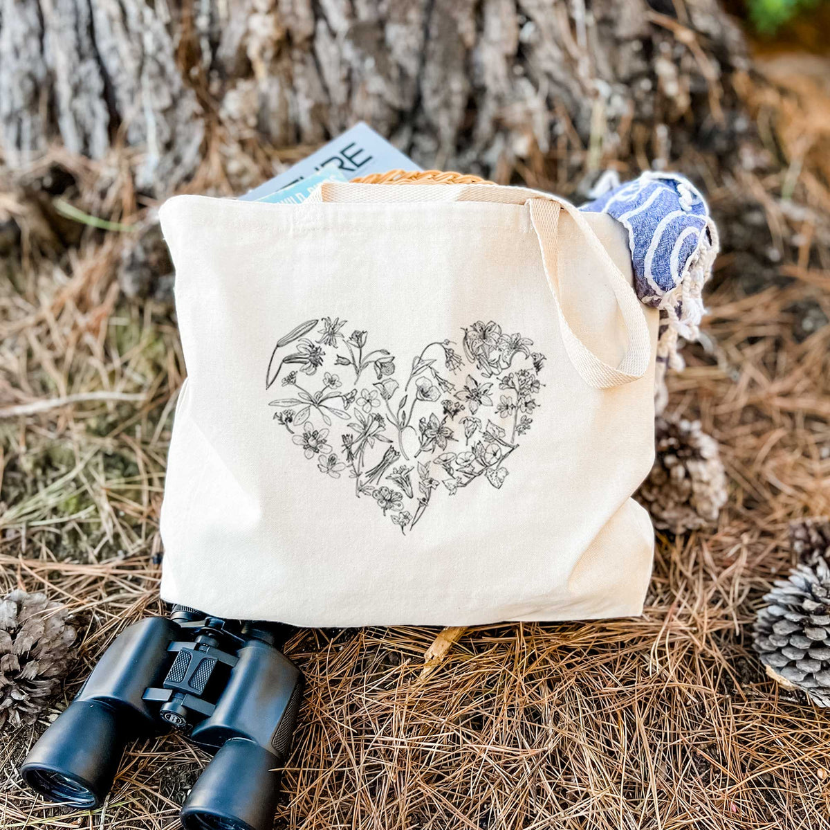 Canvas tote bag printed with a heart shape filled with black ink California mountain wildflowers, resting on dry grass beside a pair of binoculars.