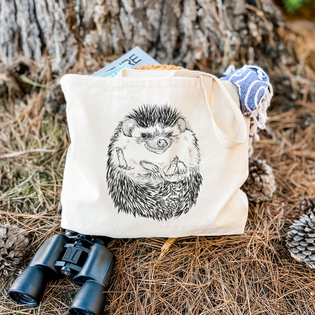 Canvas tote bag printed with a detailed black ink illustration of a hedgehog curled up and facing forward, resting on dry grass beside a pair of binoculars.