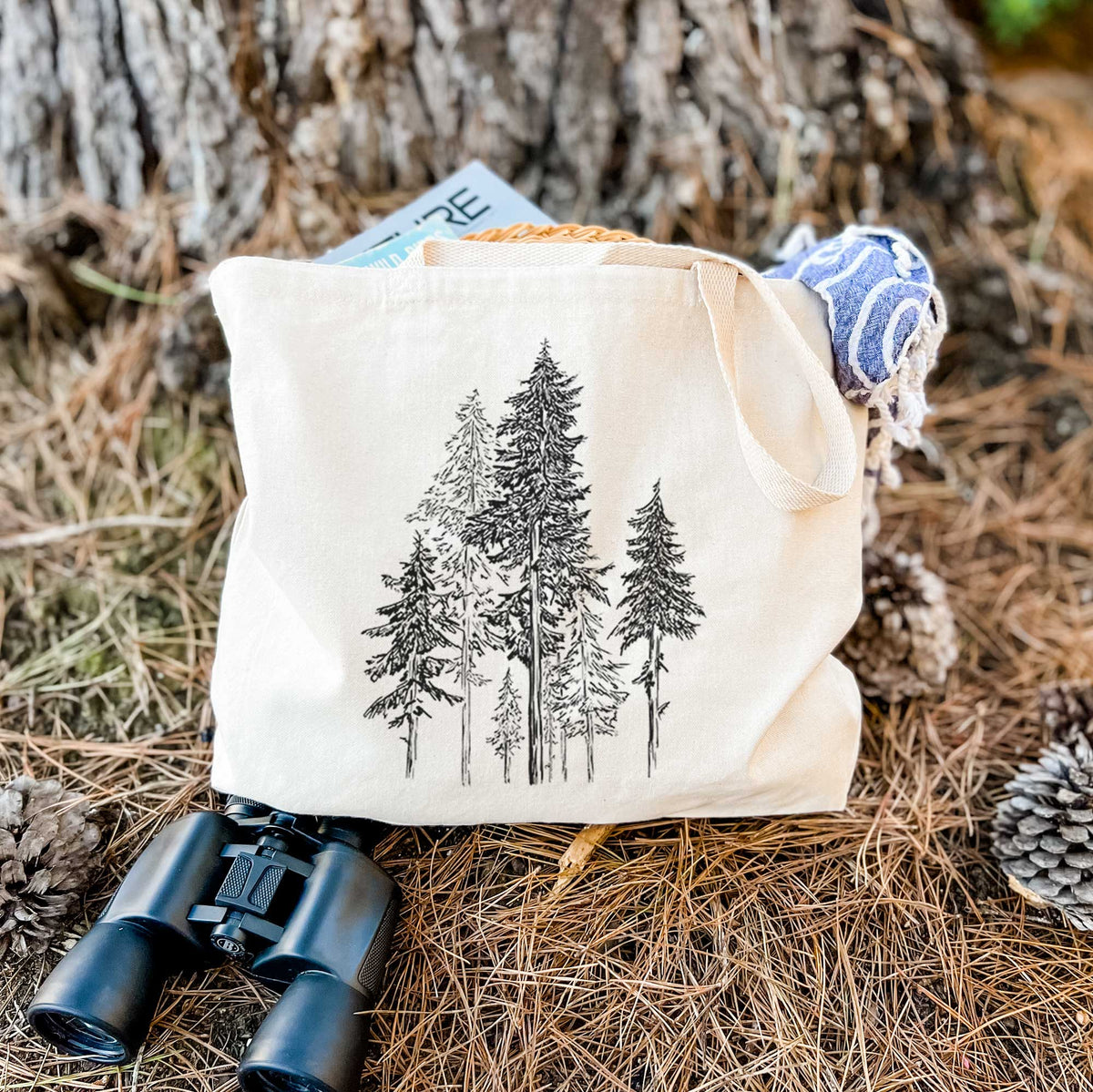 Canvas tote bag printed with a black ink illustration of a forest of tall hemlock trees, resting on dry grass beside a pair of binoculars.