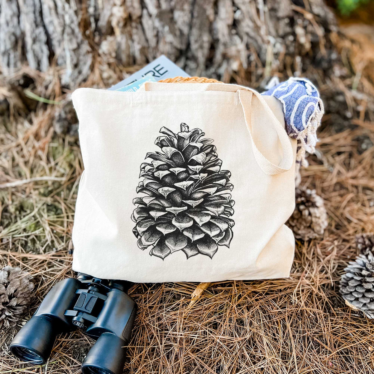 Canvas tote bag printed with a detailed black ink illustration of a large Jeffrey pine cone, resting on dry grass beside a pair of binoculars.