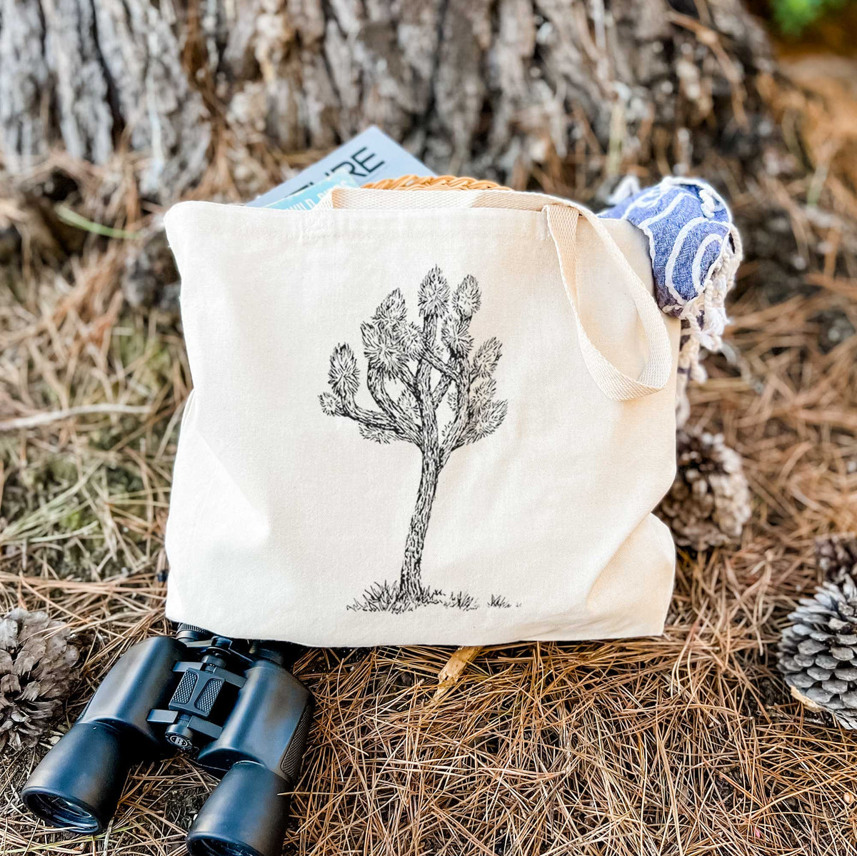 Canvas tote bag printed with a detailed black ink illustration of a Joshua tree, resting on dry grass beside a pair of binoculars.