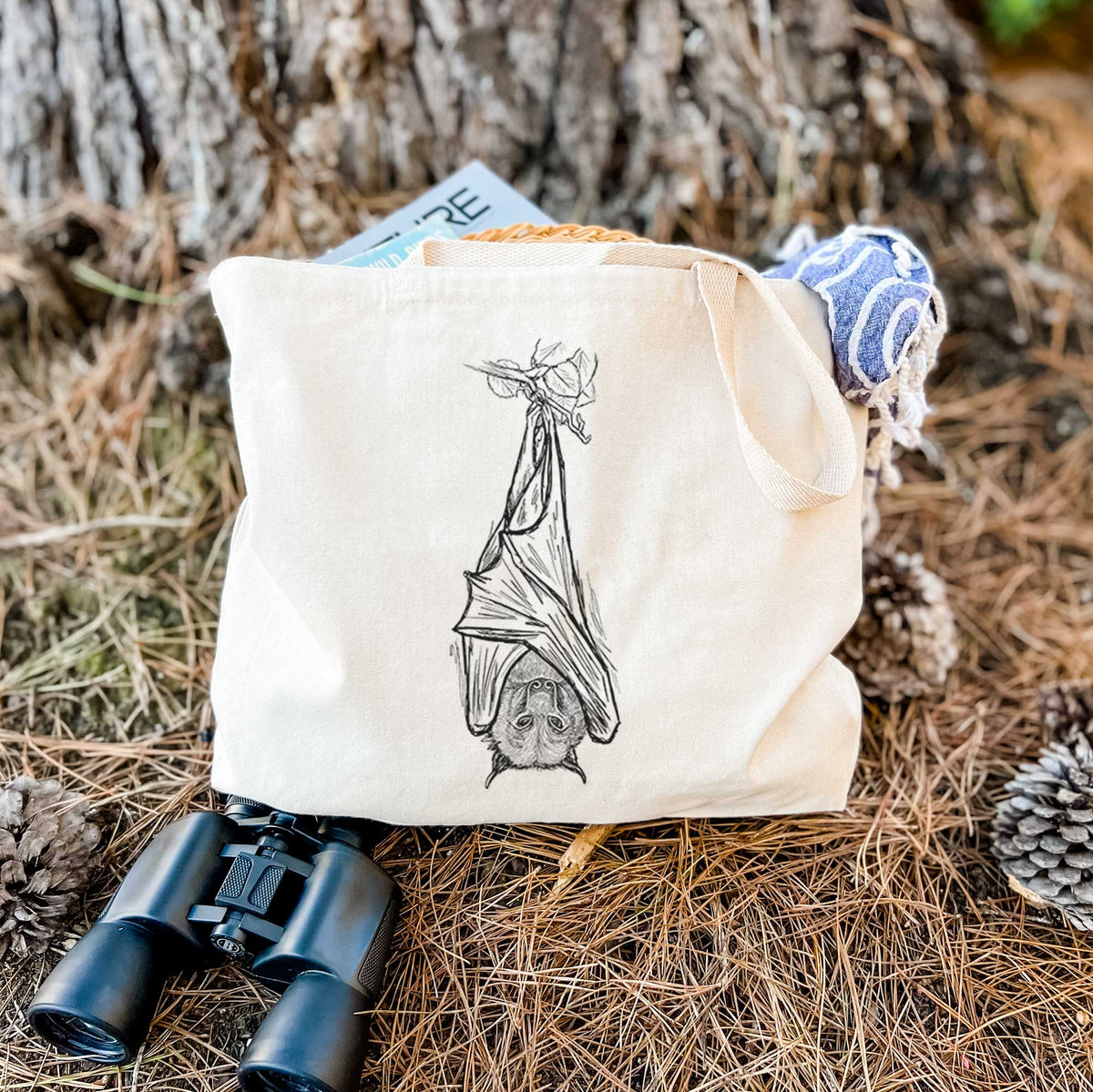 Canvas tote bag printed with a black ink illustration of a large flying fox bat hanging upside down with its wings wrapped around it, resting on dry grass beside a pair of binoculars.