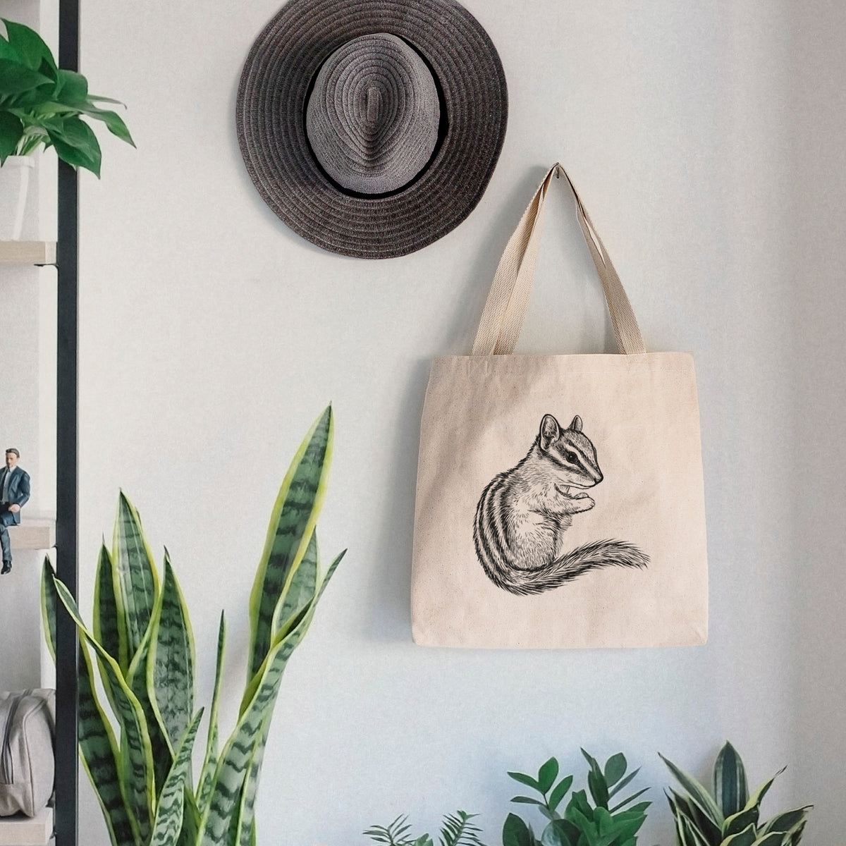 Canvas tote bag printed with a detailed black ink illustration of a least chipmunk sitting and holding food in its paws, hanging on a wall hook below a hat, next to potted plants.