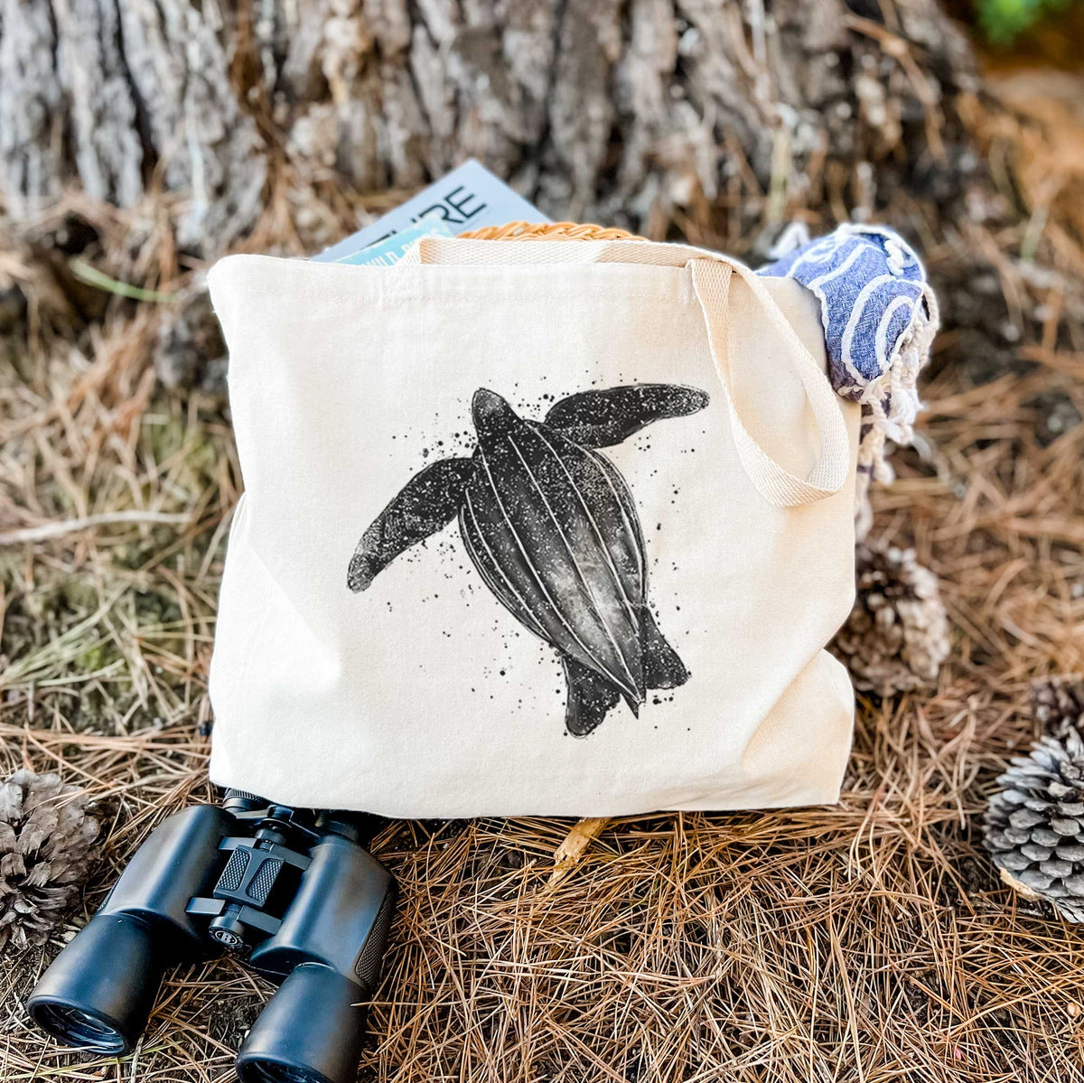 Canvas tote bag printed with a black ink illustration of a leatherback sea turtle swimming, with ink splatters, resting on dry grass beside a pair of binoculars.