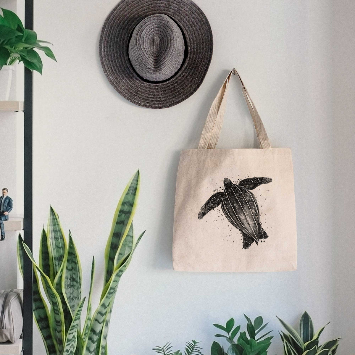 Canvas tote bag printed with a black ink illustration of a leatherback sea turtle swimming, with ink splatters, hanging on a wall hook below a hat, next to potted plants.