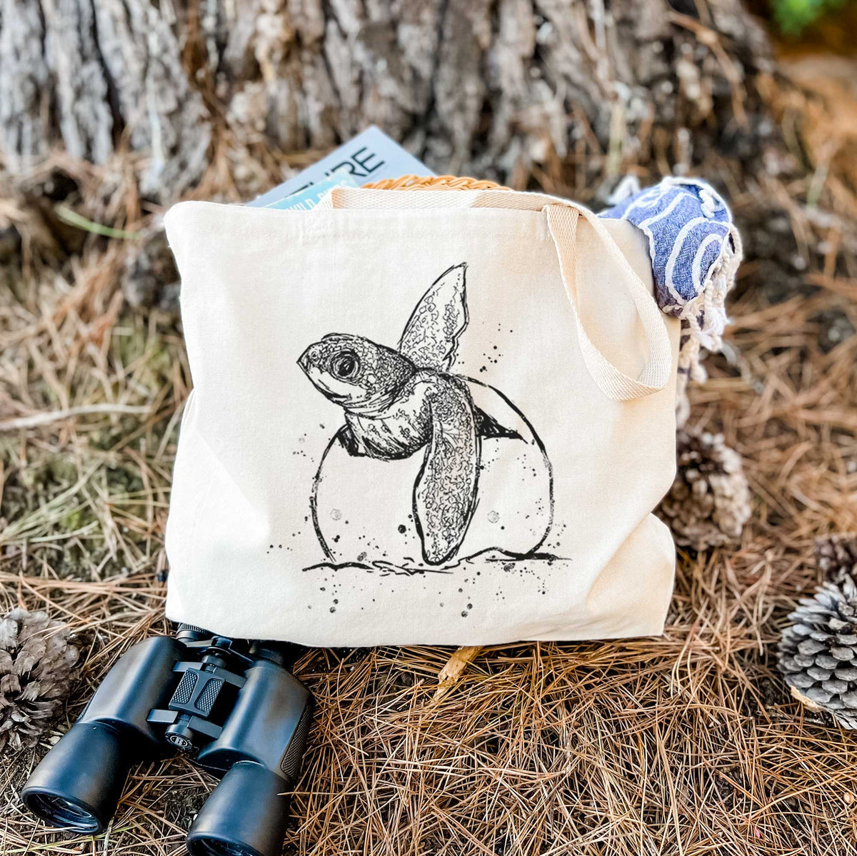 Canvas tote bag printed with a black ink illustration of a leatherback sea turtle hatchling emerging from its egg, resting on dry grass beside a pair of binoculars.