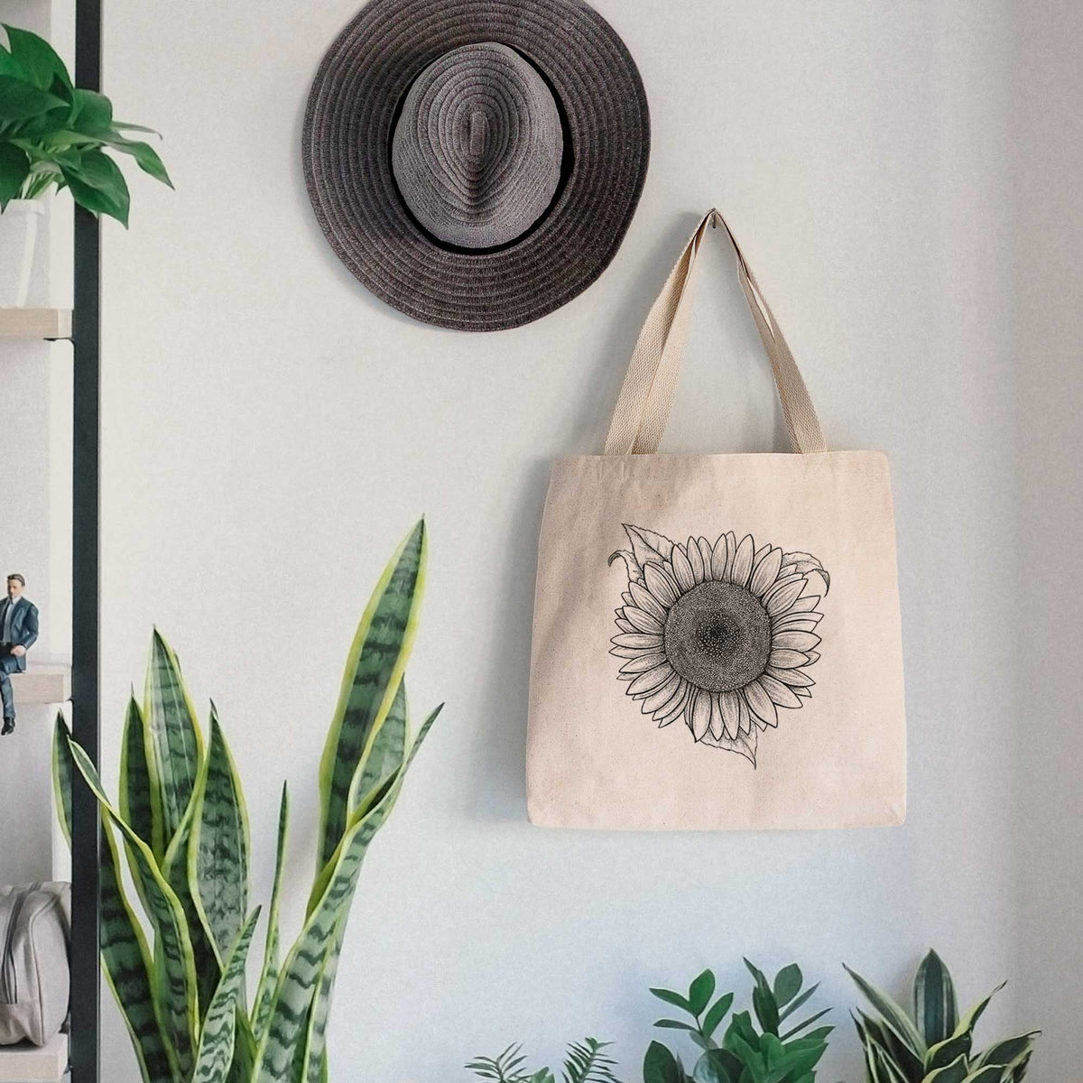 Canvas tote bag printed with a detailed black ink line drawing of a Lemon Queen sunflower in bloom, hanging on a wall hook below a hat, next to potted plants.