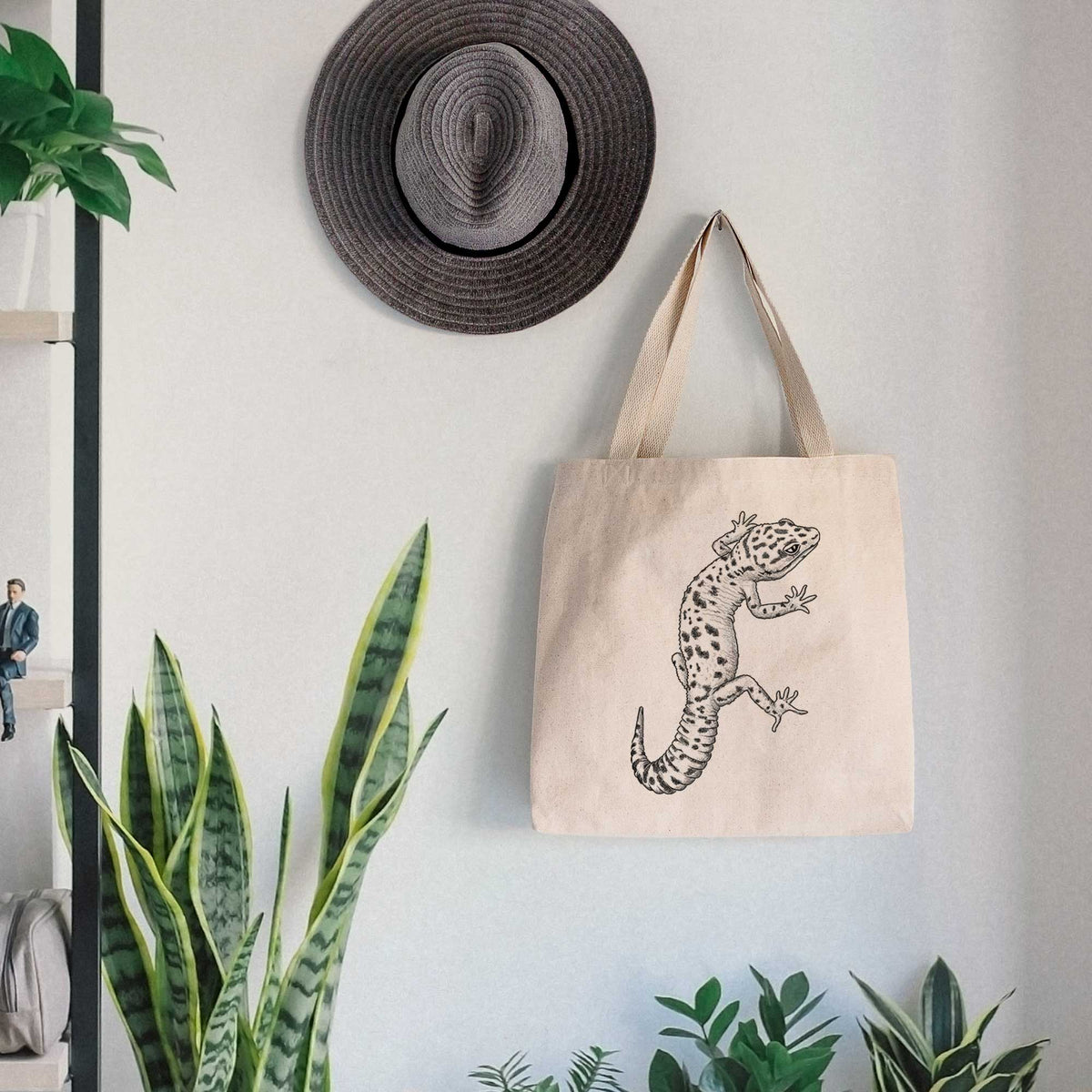 Canvas tote bag printed with a detailed black ink illustration of a spotted leopard gecko climbing, hanging on a wall hook below a hat, next to potted plants.