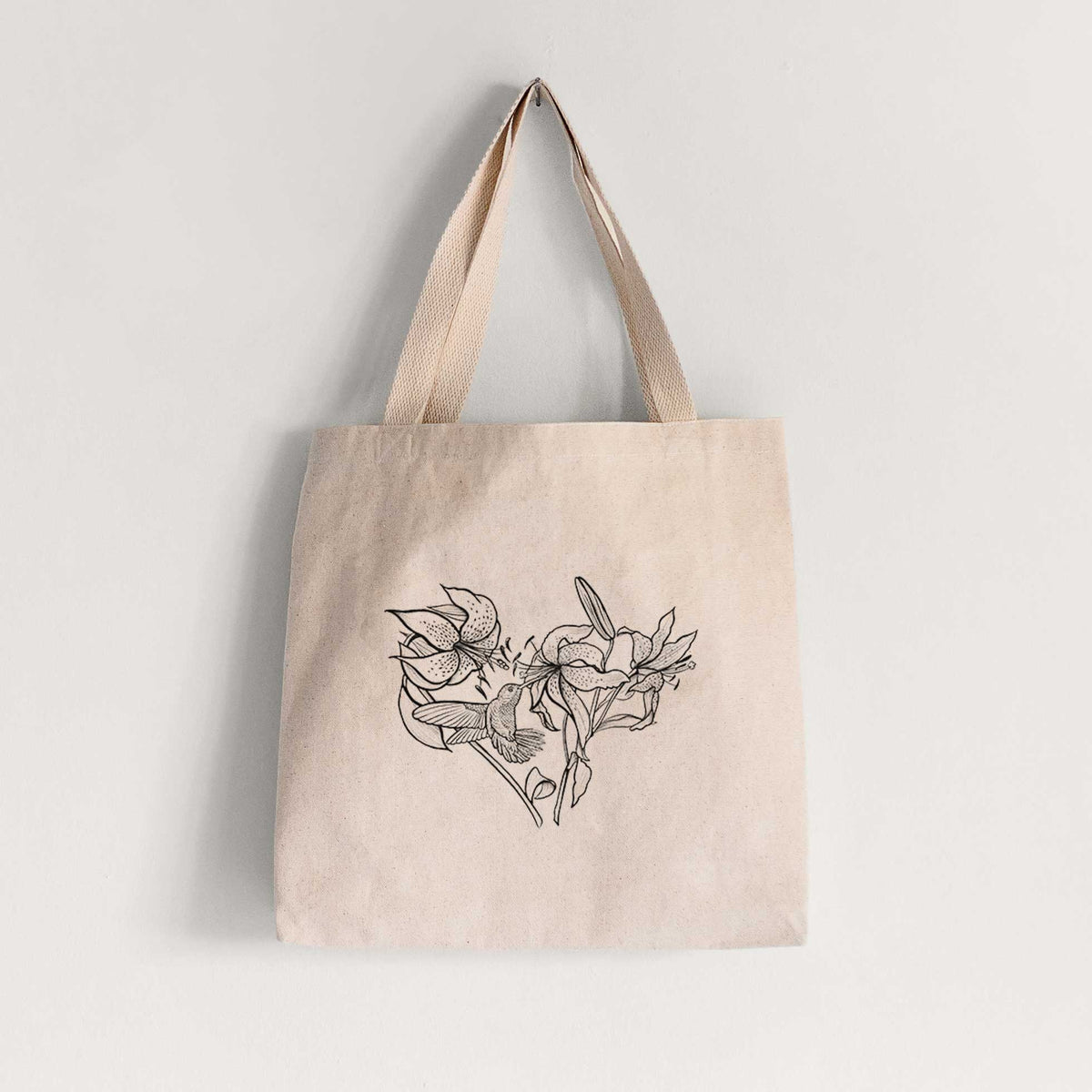 Natural canvas tote bag printed with a black ink line drawing of lilies and a hummingbird arranged in a heart shape.