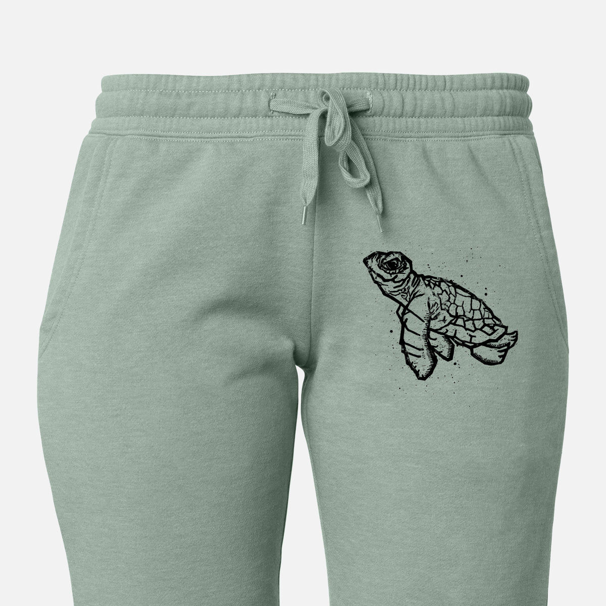 Front of sage green Cali Wave sweatpants, printed with a black linocut-style illustration of a baby sea turtle with a patterned shell and ink splatters on the leg.