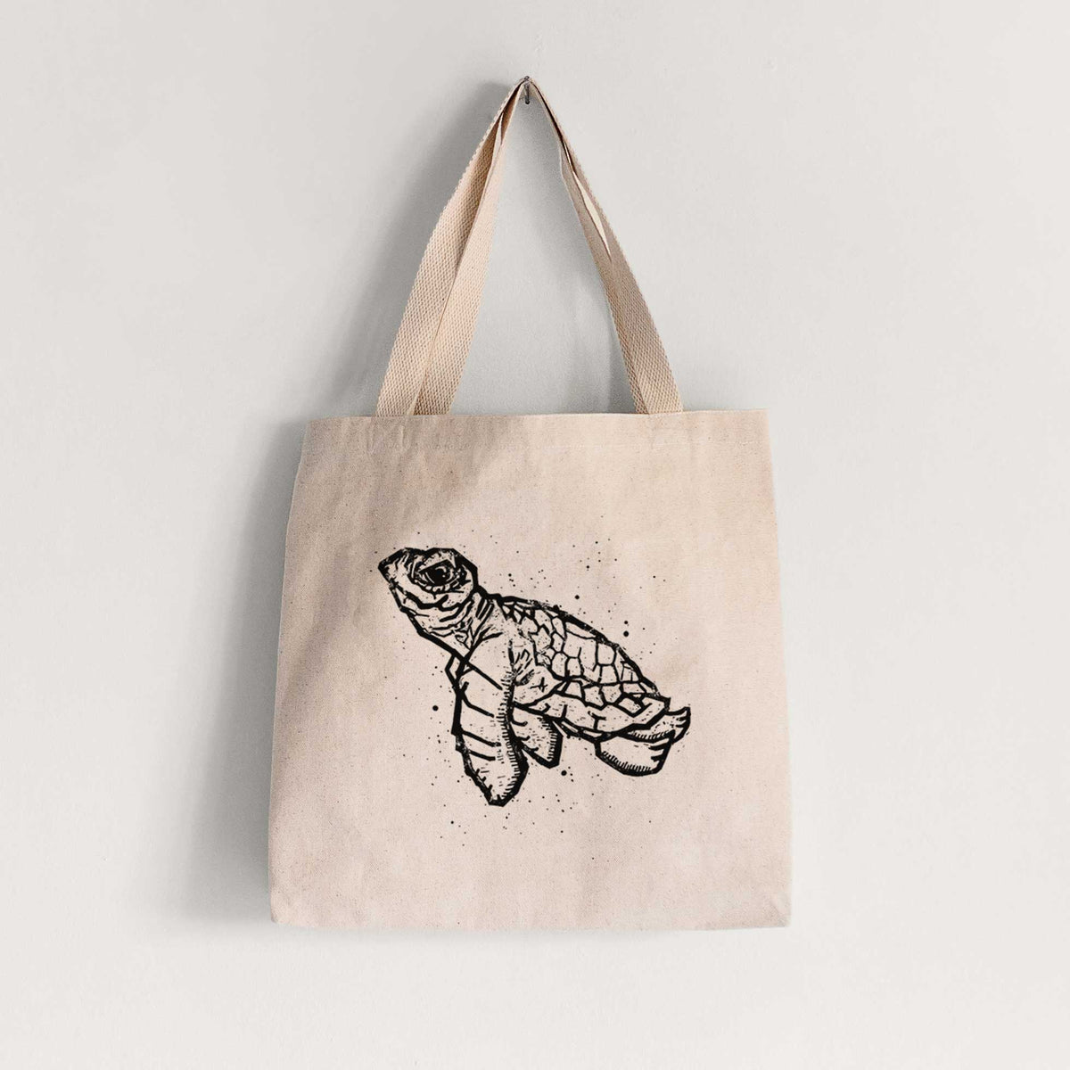 Natural canvas tote bag printed with a black linocut-style illustration of a baby sea turtle with a patterned shell and ink splatters.