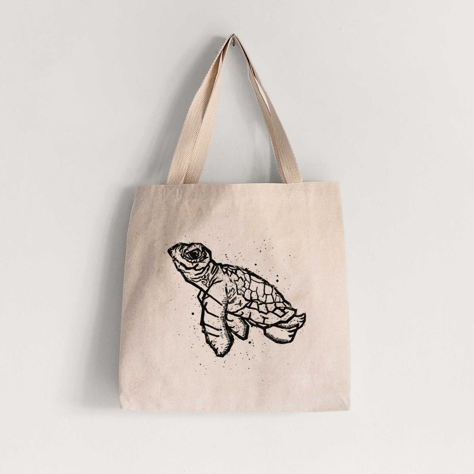 Natural canvas tote bag printed with a black linocut-style illustration of a baby sea turtle with a patterned shell and ink splatters.