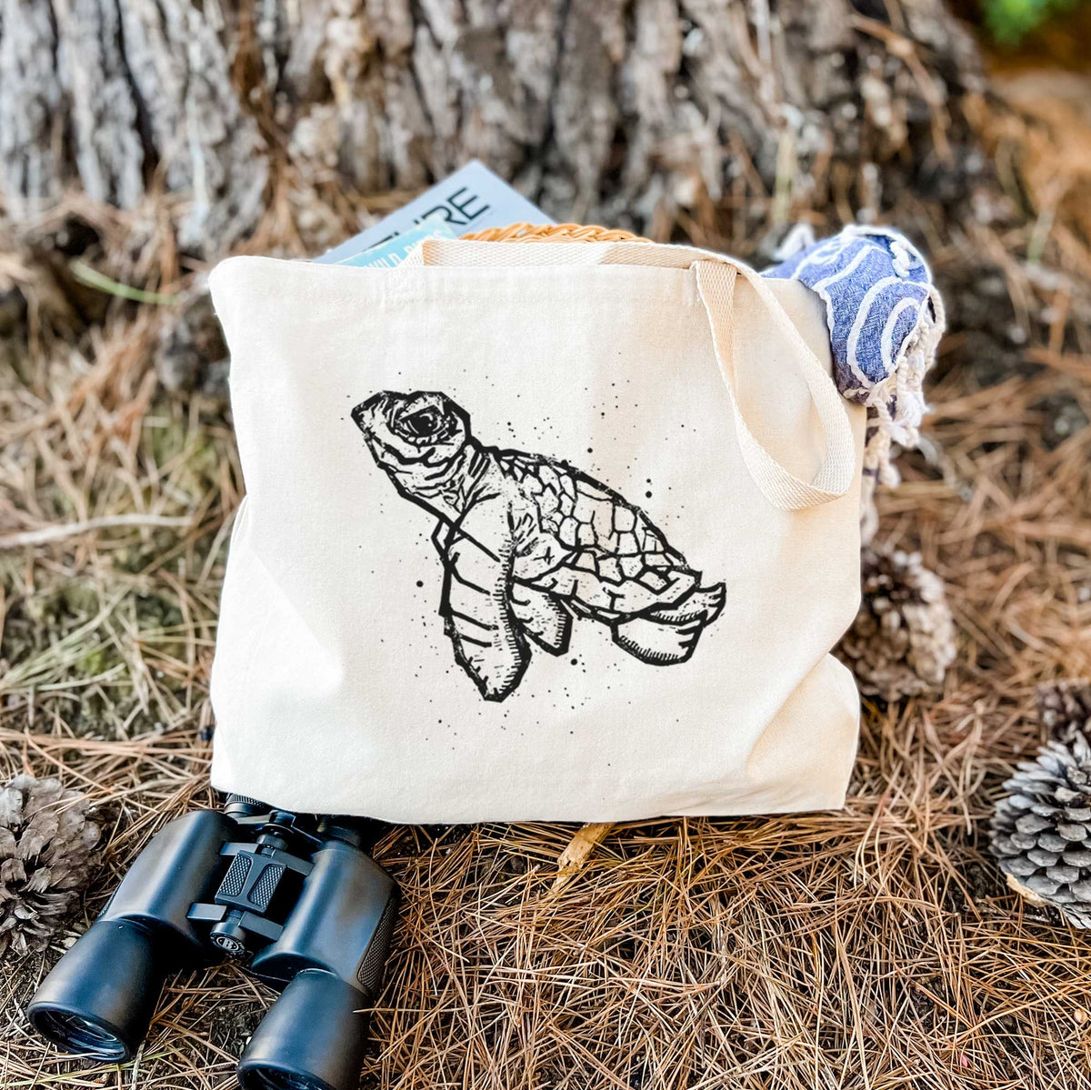 Canvas tote bag printed with a black linocut-style illustration of a baby sea turtle with a patterned shell and ink splatters, resting on dry grass beside a pair of binoculars.