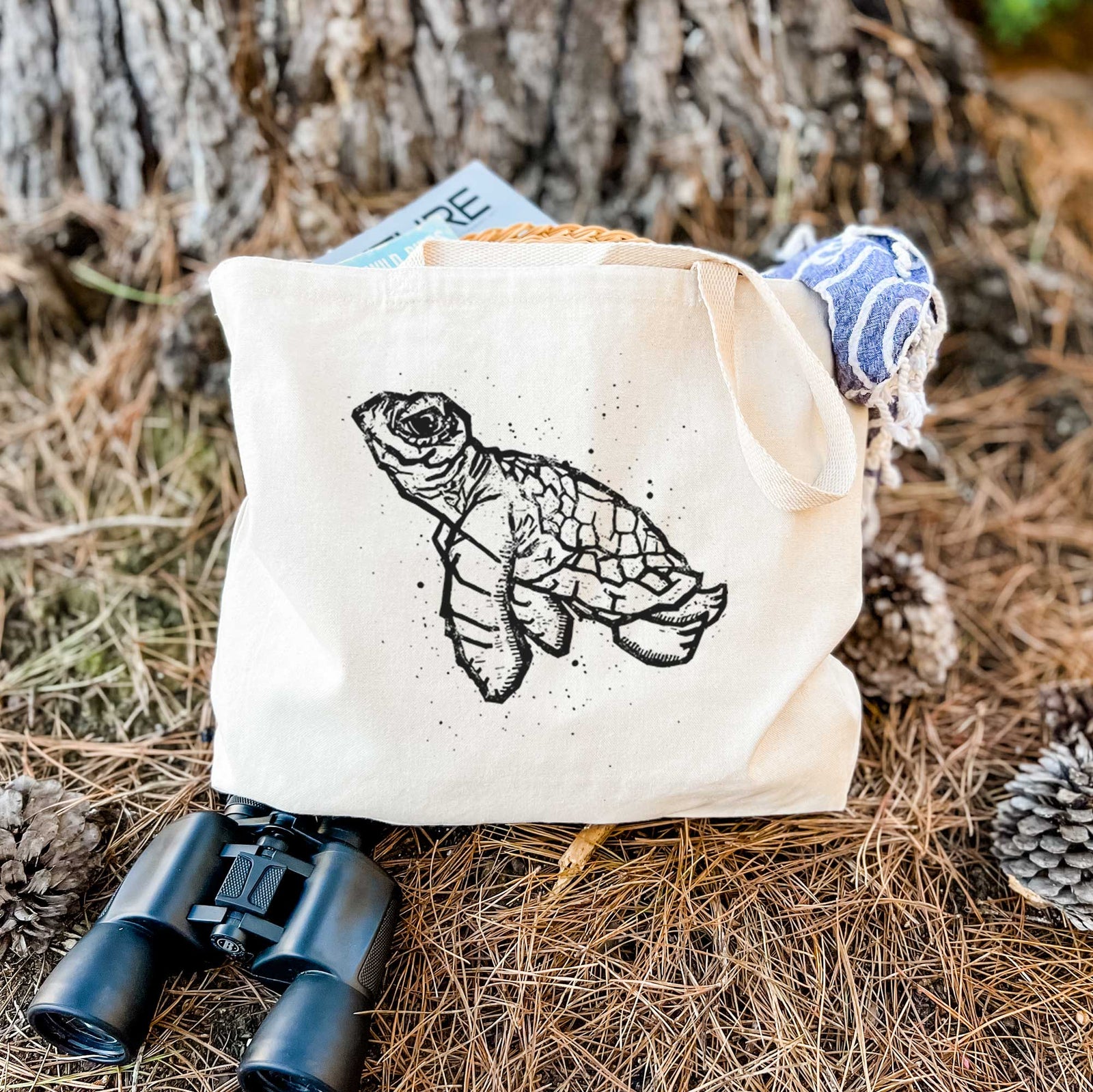 Natural canvas tote bag printed with a black linocut-style illustration of a baby sea turtle with a patterned shell and ink splatters.