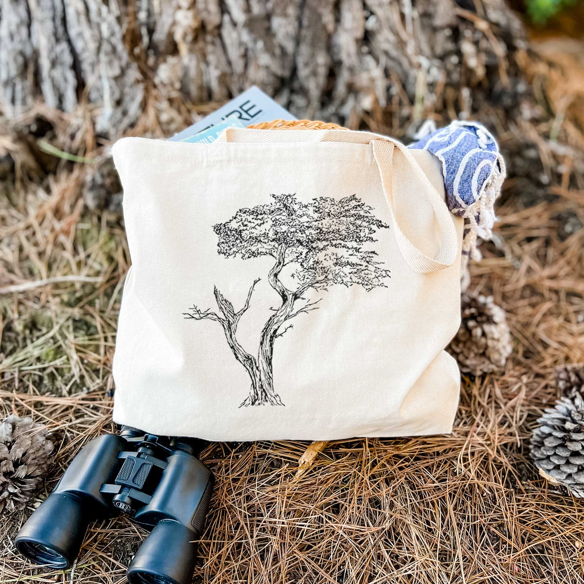 Canvas tote bag printed with a black ink illustration of the windswept Lone Cypress tree on a rocky outcrop, resting on dry grass beside a pair of binoculars.
