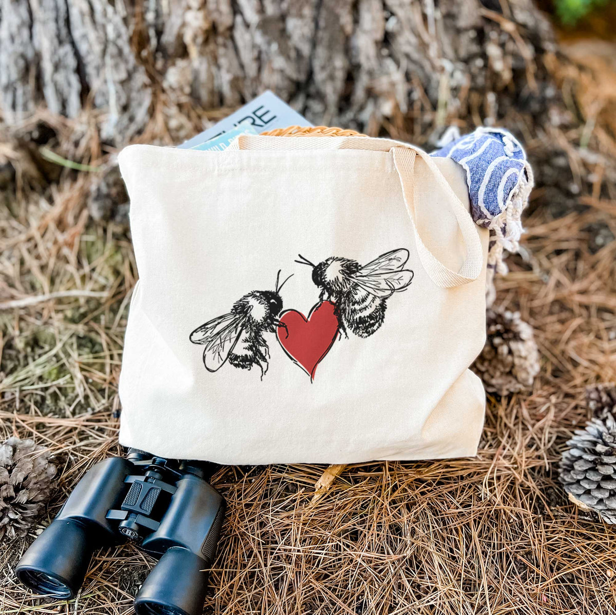 Canvas tote bag printed with a black ink illustration of two bees facing each other with a red heart between them, resting on dry grass beside a pair of binoculars.