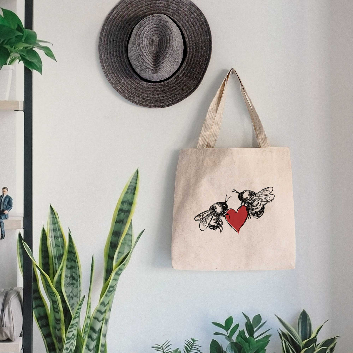 Canvas tote bag printed with a black ink illustration of two bees facing each other with a red heart between them, hanging on a wall hook below a hat, next to potted plants.