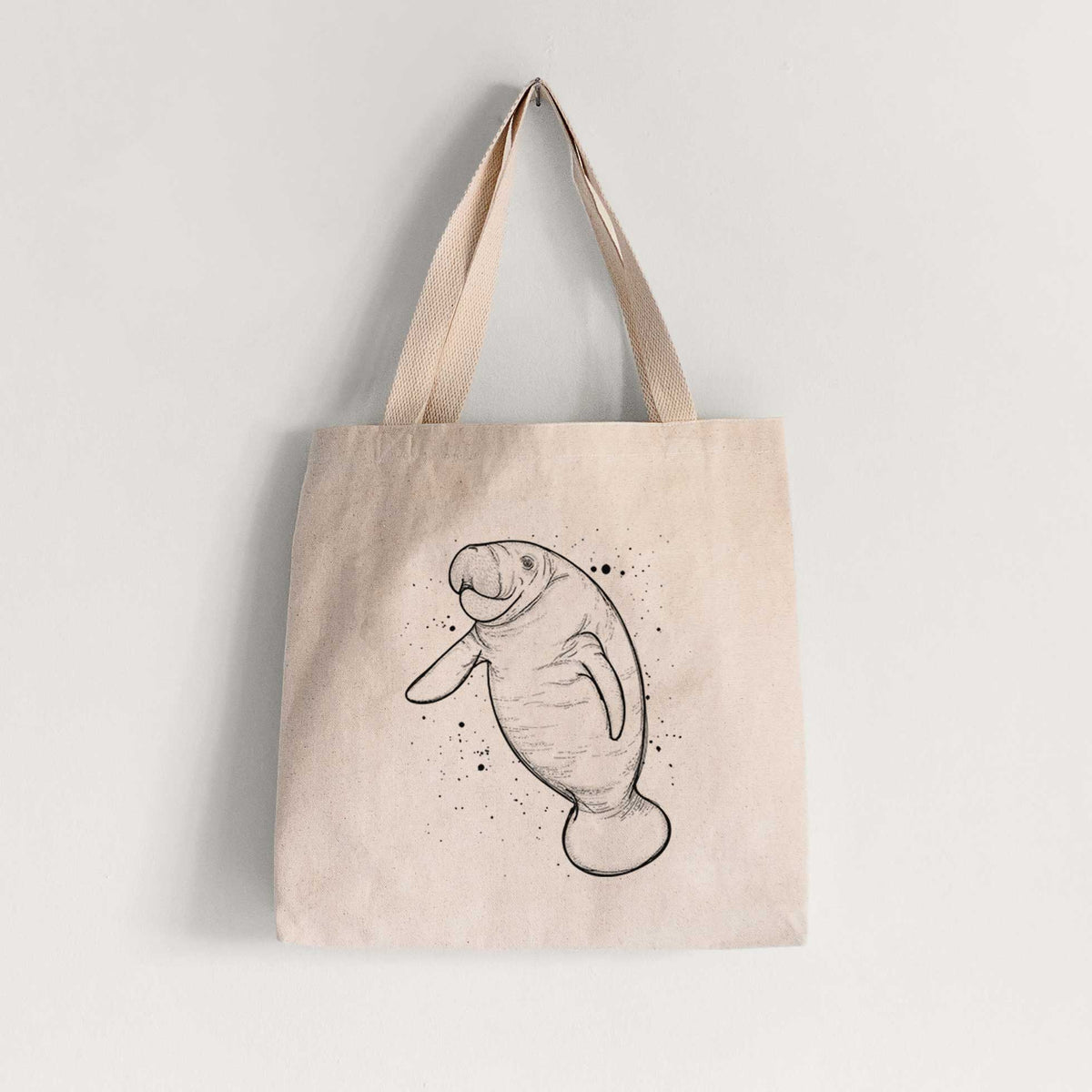 Natural canvas tote bag printed with a black ink line drawing of a manatee swimming, with ink splatters.