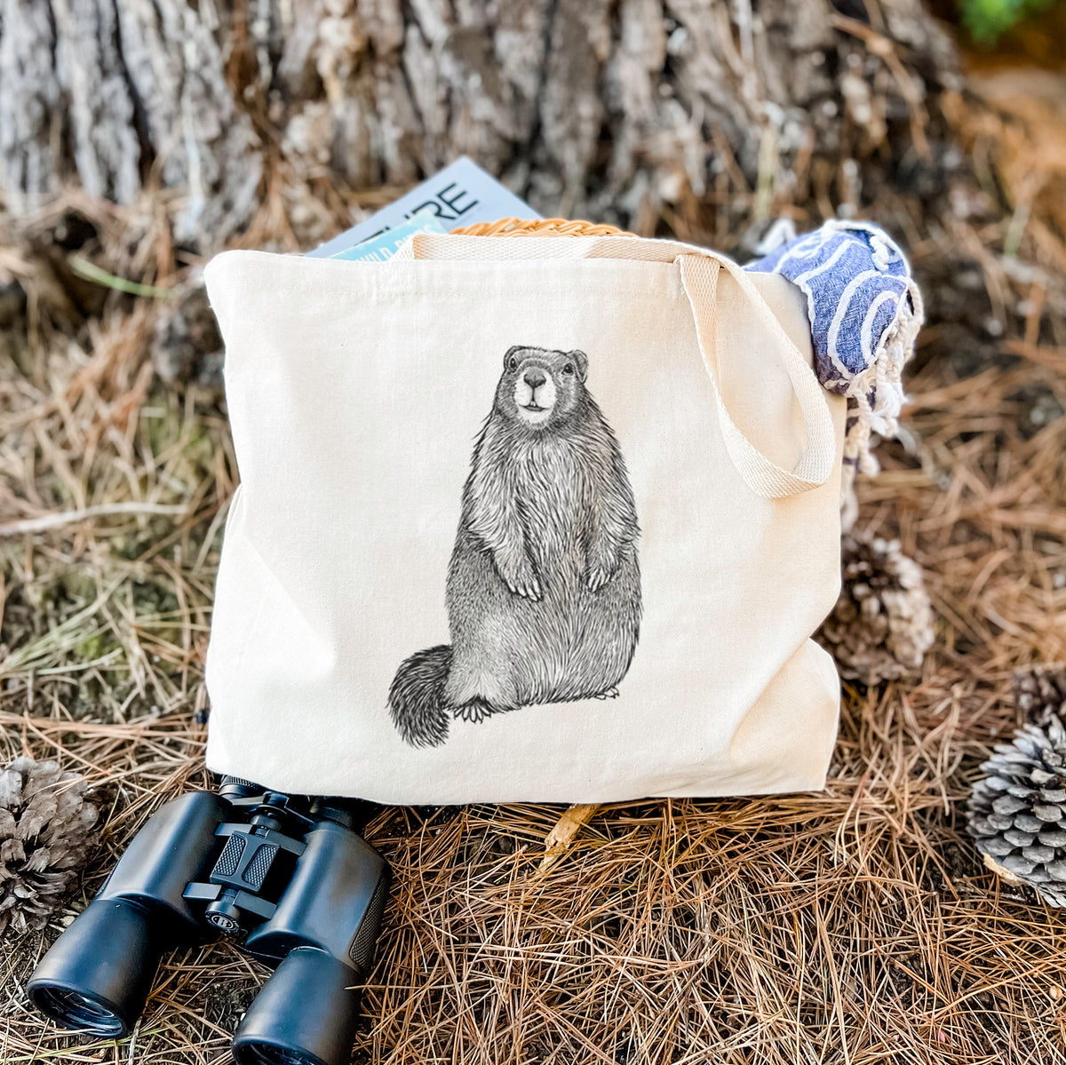 Canvas tote bag printed with a detailed black ink illustration of a marmot standing upright, resting on dry grass beside a pair of binoculars.