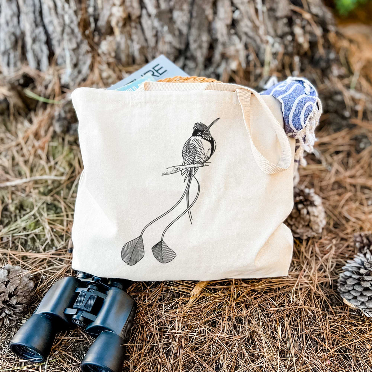 Canvas tote bag printed with a black ink illustration of a marvelous spatuletail hummingbird perched, with its two long racket-tipped tail feathers, resting on dry grass beside a pair of binoculars.