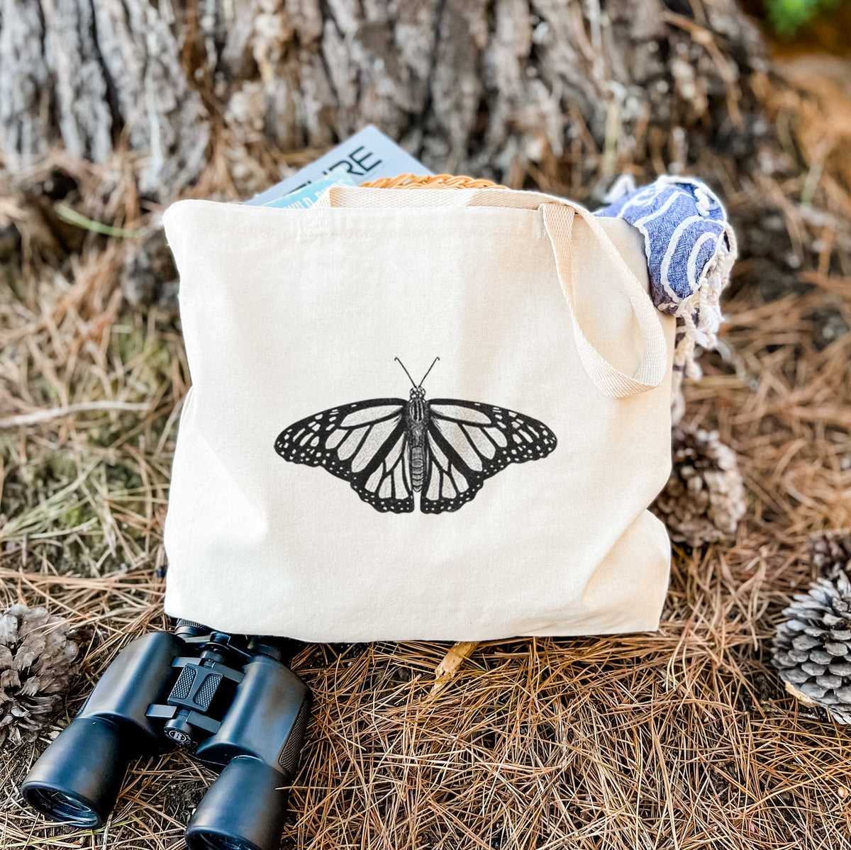 Canvas tote bag printed with a detailed black ink illustration of a monarch butterfly with its wings spread, resting on dry grass beside a pair of binoculars.