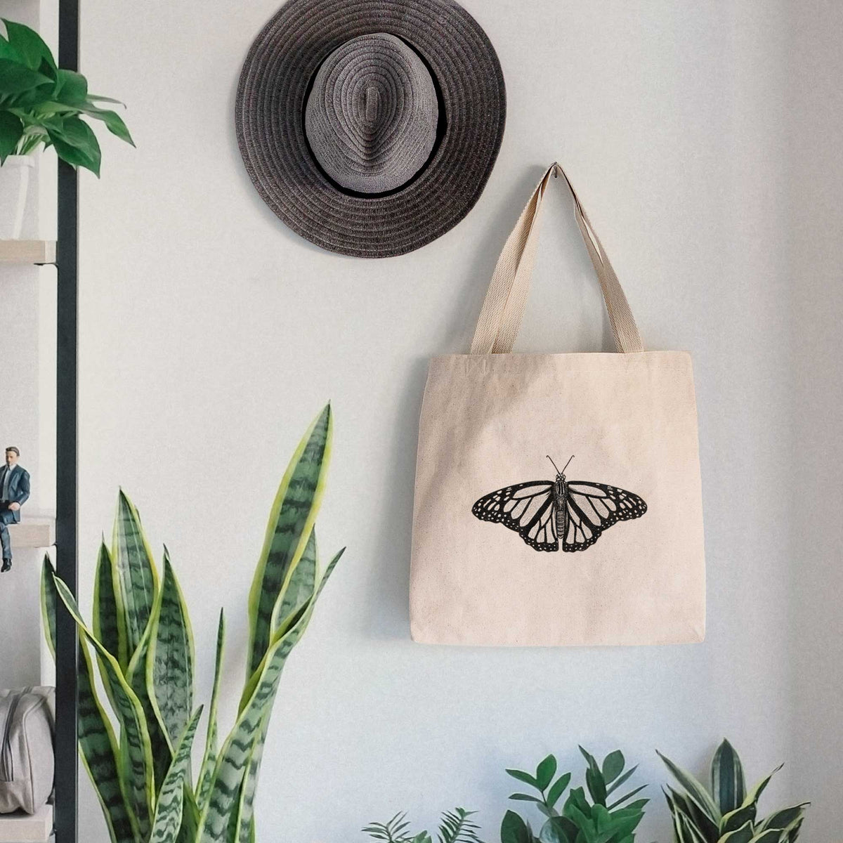 Canvas tote bag printed with a detailed black ink illustration of a monarch butterfly with its wings spread, hanging on a wall hook below a hat, next to potted plants.