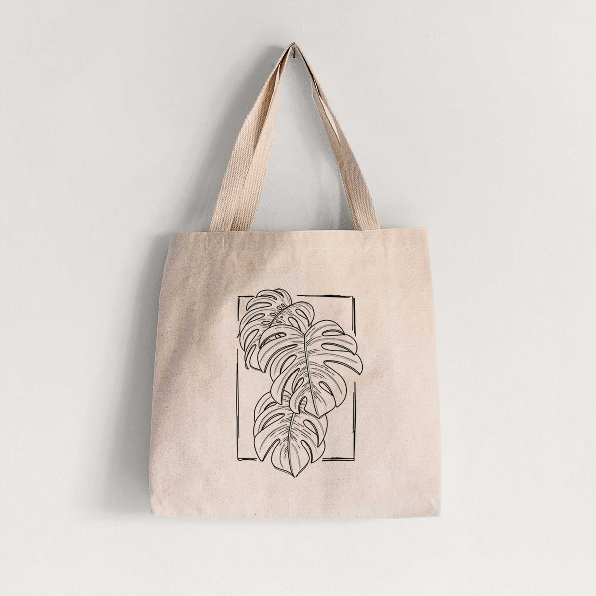 Natural canvas tote bag printed with a black ink line drawing of Monstera deliciosa leaves inside a rectangle outline.