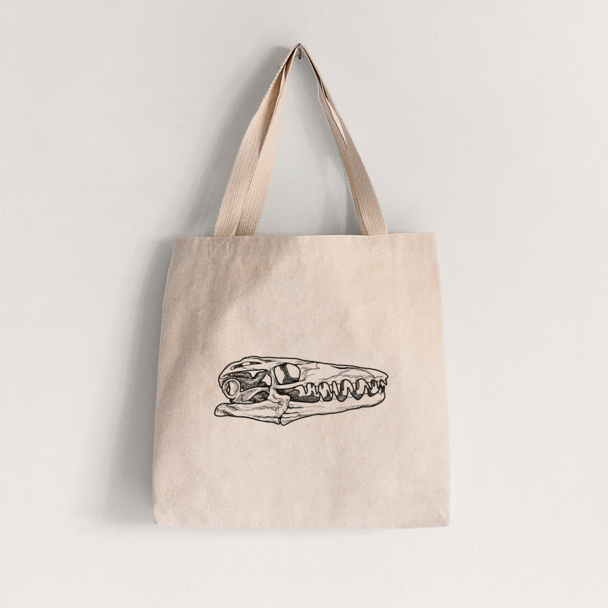 Natural canvas tote bag printed with a detailed black ink illustration of a Mosasaur skull.