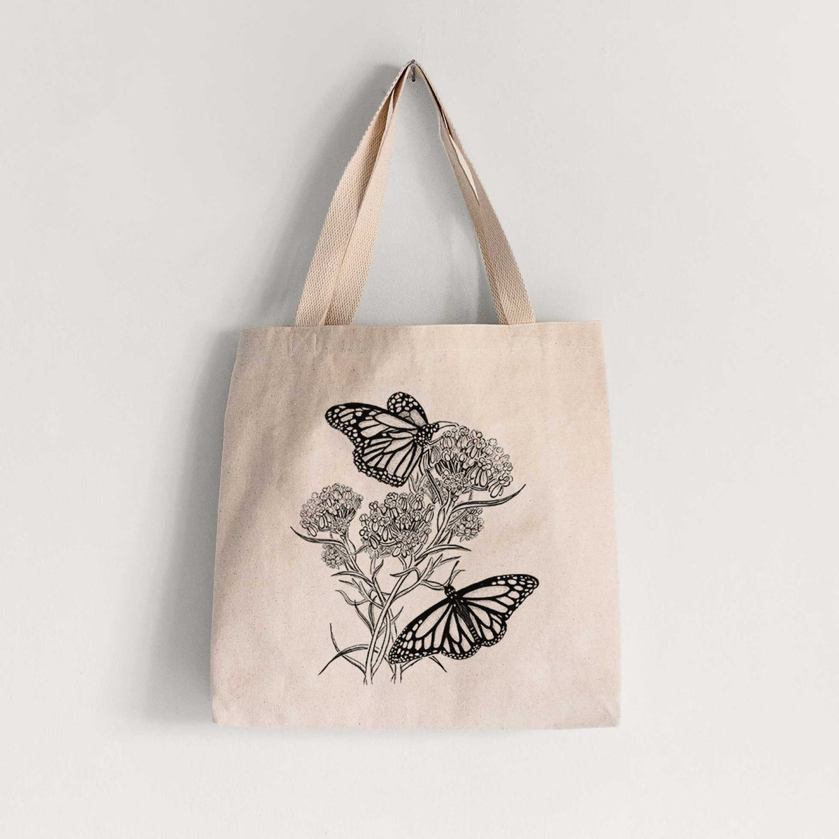 Natural canvas tote bag printed with a black ink illustration of two monarch butterflies on flowering milkweed.