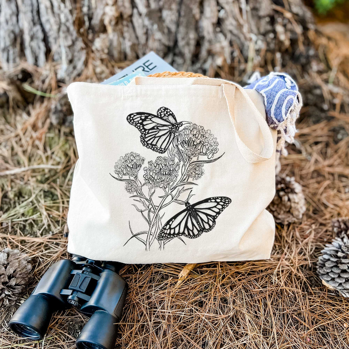 Canvas tote bag printed with a black ink illustration of two monarch butterflies on flowering milkweed, resting on dry grass beside a pair of binoculars.