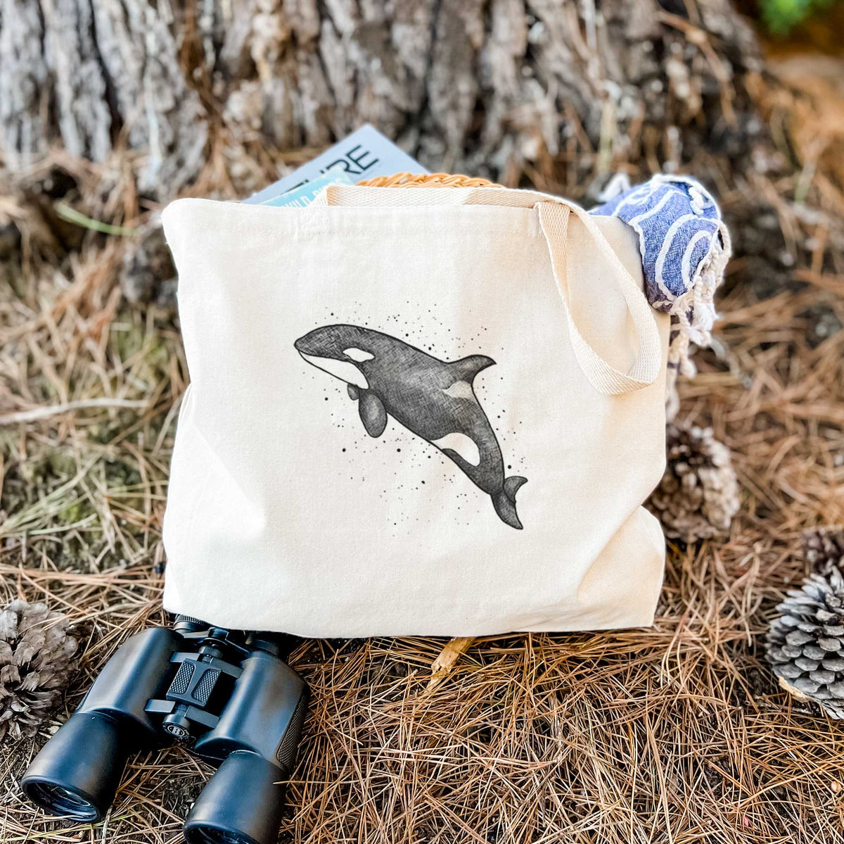 Canvas tote bag printed with a black ink illustration of an orca leaping, with ink splatters, resting on dry grass beside a pair of binoculars.