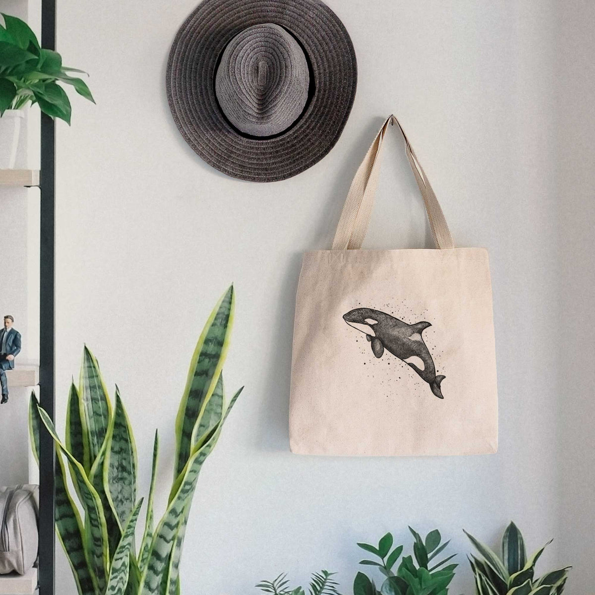 Canvas tote bag printed with a black ink illustration of an orca leaping, with ink splatters, hanging on a wall hook below a hat, next to potted plants.
