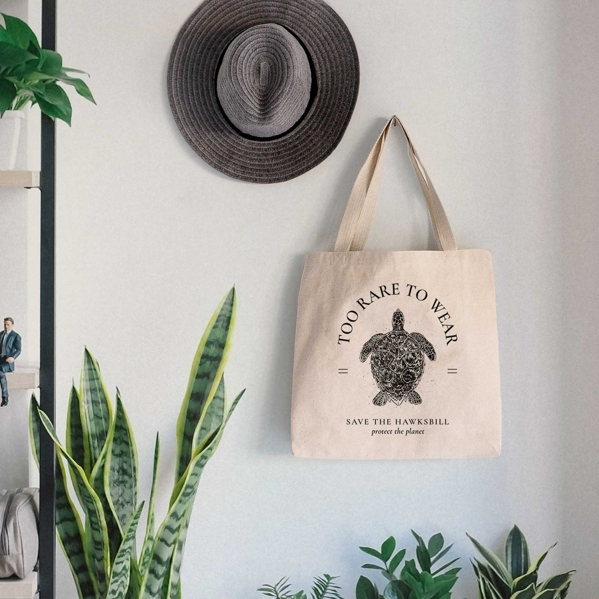 Canvas tote bag printed with a black ink hawksbill sea turtle framed by the words Too Rare to Wear, Save the Hawksbill, hanging on a wall hook below a hat, next to potted plants.
