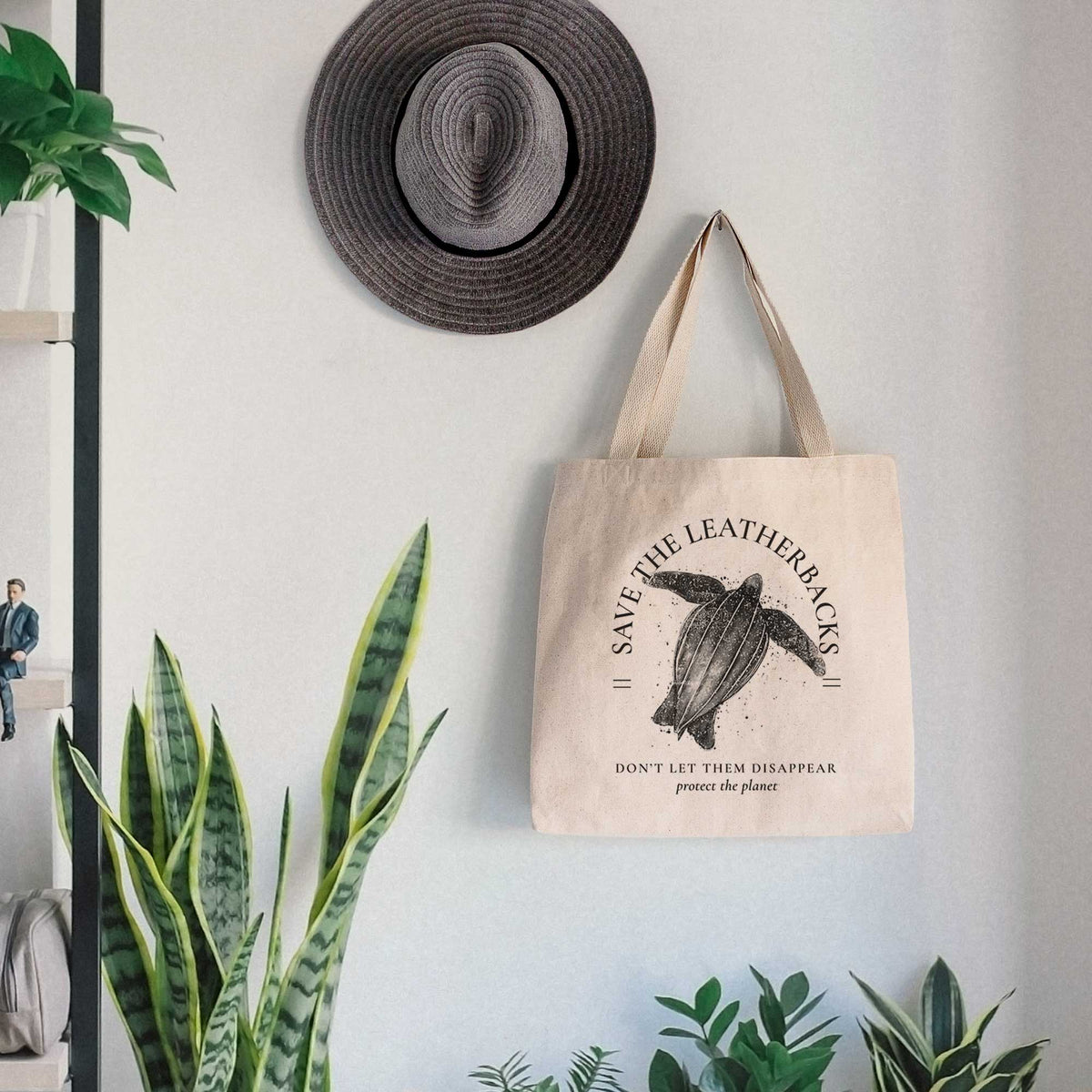 Canvas tote bag printed with a black ink leatherback sea turtle framed by the words Save the Leatherbacks, Don't Let Them Disappear, hanging on a wall hook below a hat, next to potted plants.