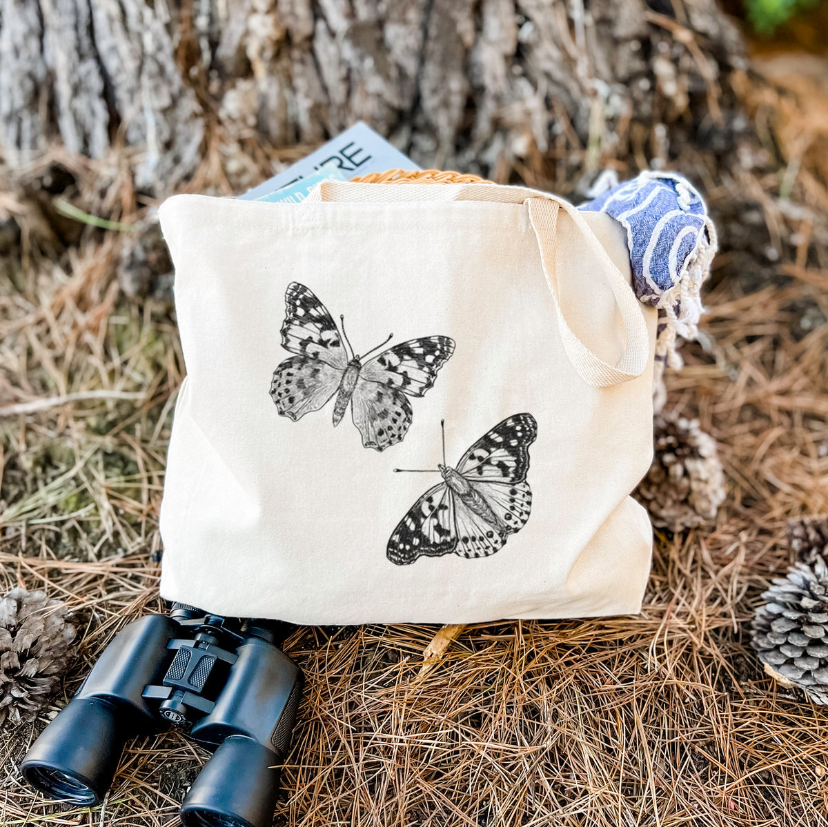 Canvas tote bag printed with a detailed black ink illustration of two painted lady butterflies, resting on dry grass beside a pair of binoculars.