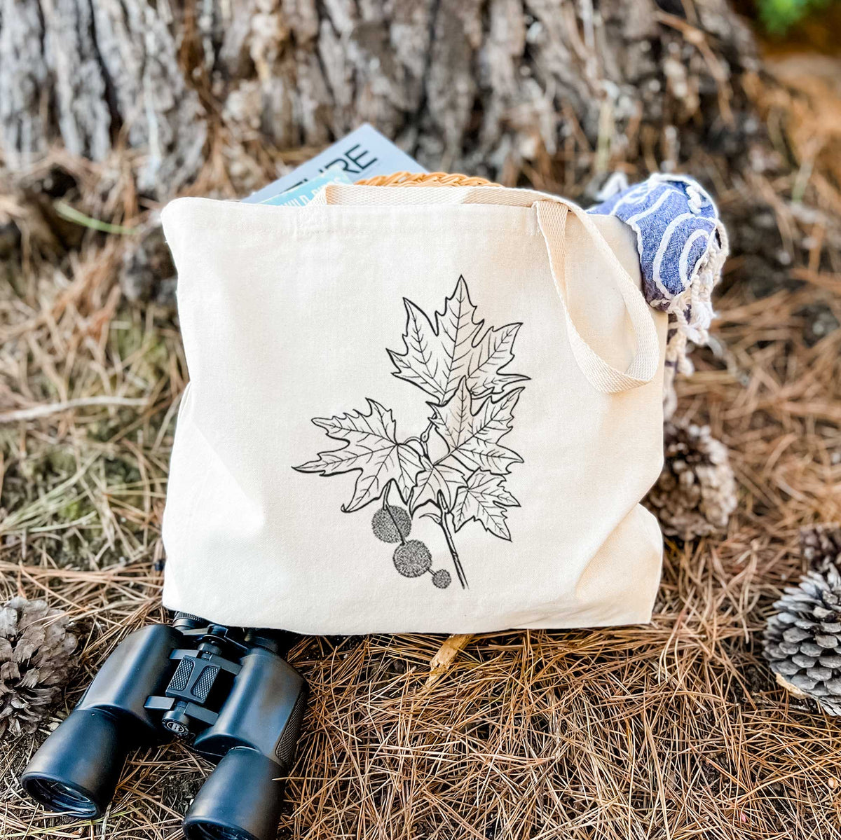 Canvas tote bag printed with a black ink illustration of an oriental plane tree branch with lobed leaves and round seed balls, resting on dry grass beside a pair of binoculars.