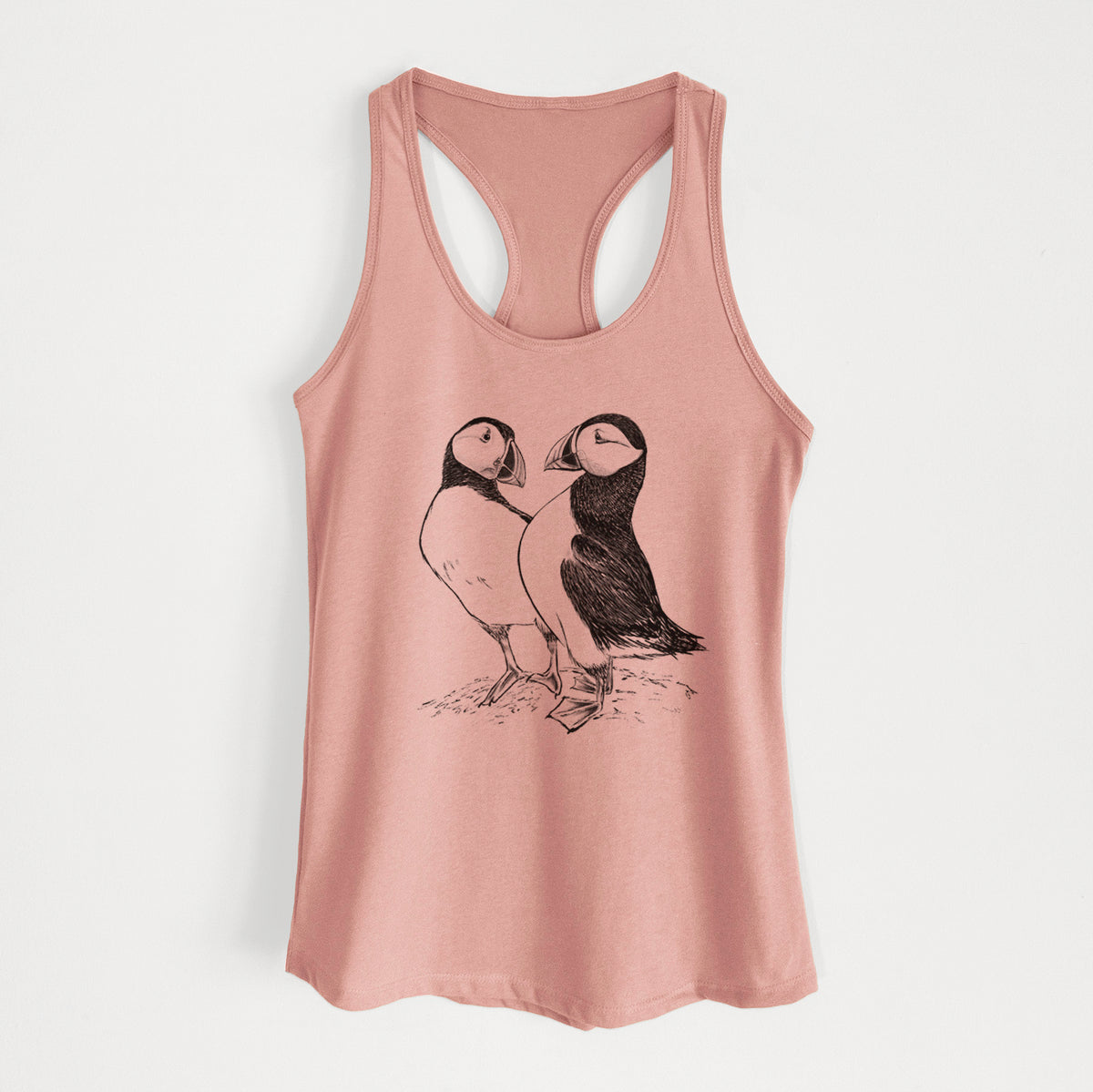 Desert pink women's racerback tank top laid flat, printed with a detailed black ink illustration of two Atlantic puffins standing side by side.