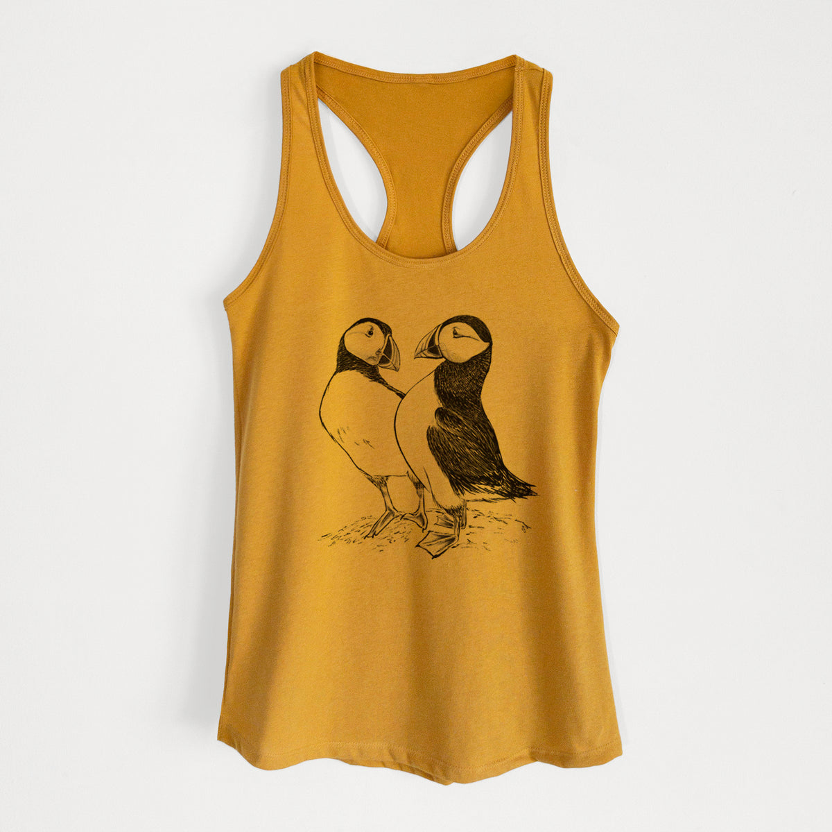 Gold women's racerback tank top laid flat, printed with a detailed black ink illustration of two Atlantic puffins standing side by side.
