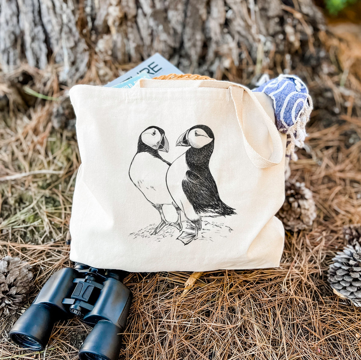 Canvas tote bag printed with a detailed black ink illustration of two Atlantic puffins standing side by side, resting on dry grass beside a pair of binoculars.
