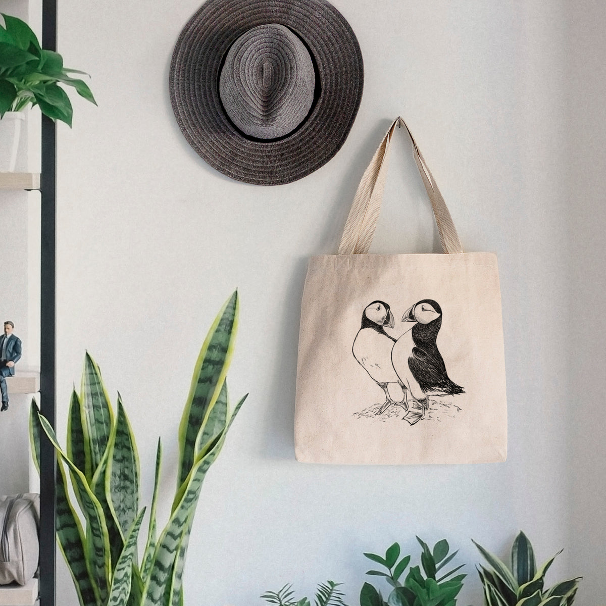 Canvas tote bag printed with a detailed black ink illustration of two Atlantic puffins standing side by side, hanging on a wall hook below a hat, next to potted plants.