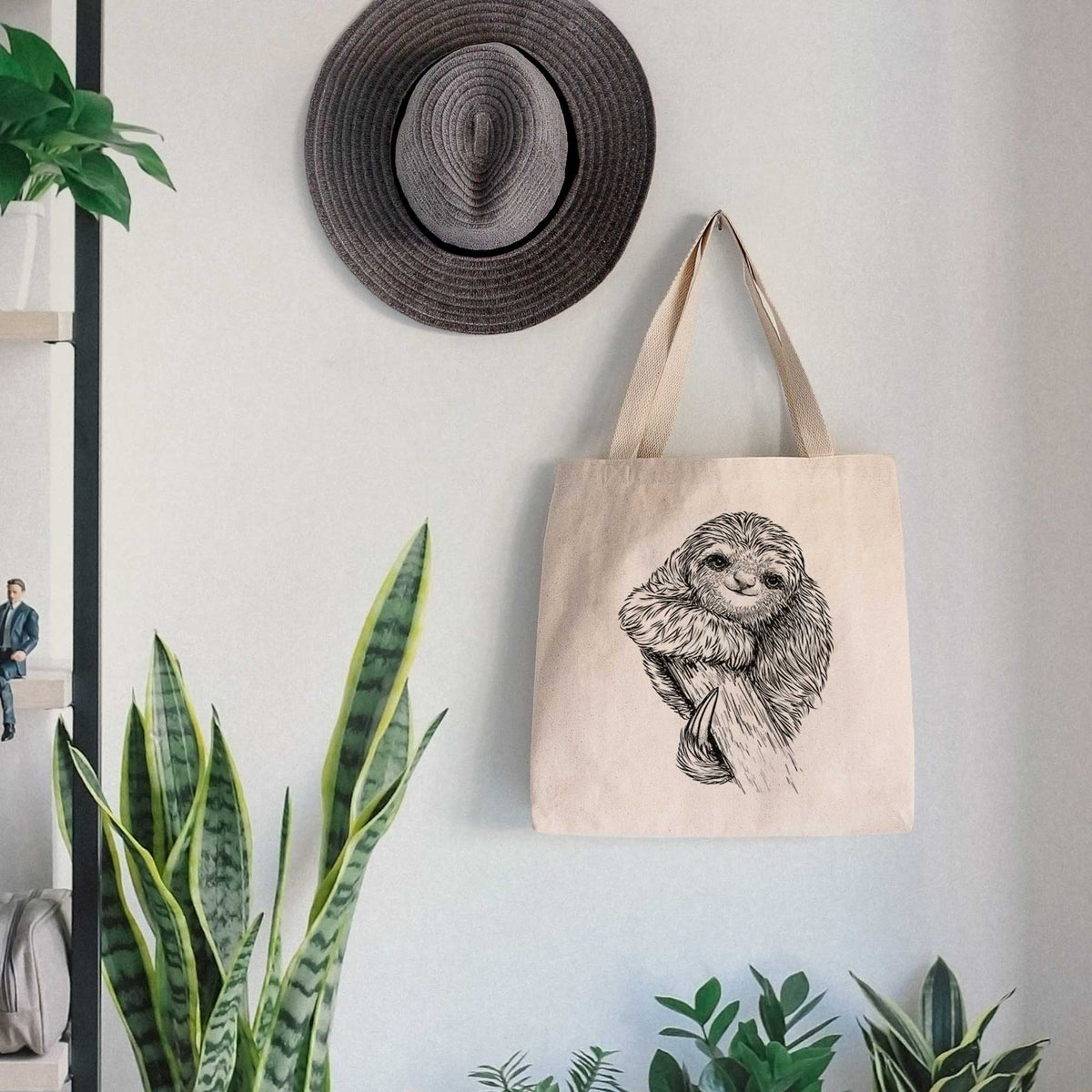 Canvas tote bag printed with a detailed black ink illustration of a smiling pygmy three-toed sloth hanging from a branch, hanging on a wall hook below a hat, next to potted plants.